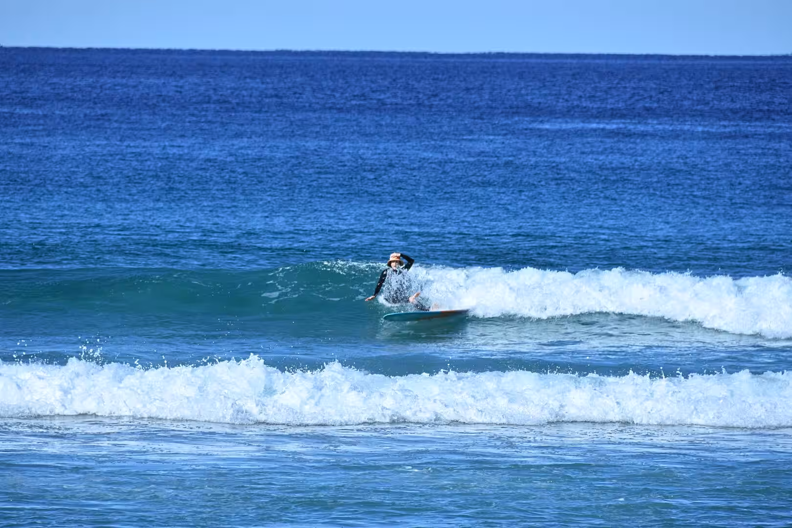 A surfer in a wetsuit rides a low breaking wave over deep blue water and white foam.