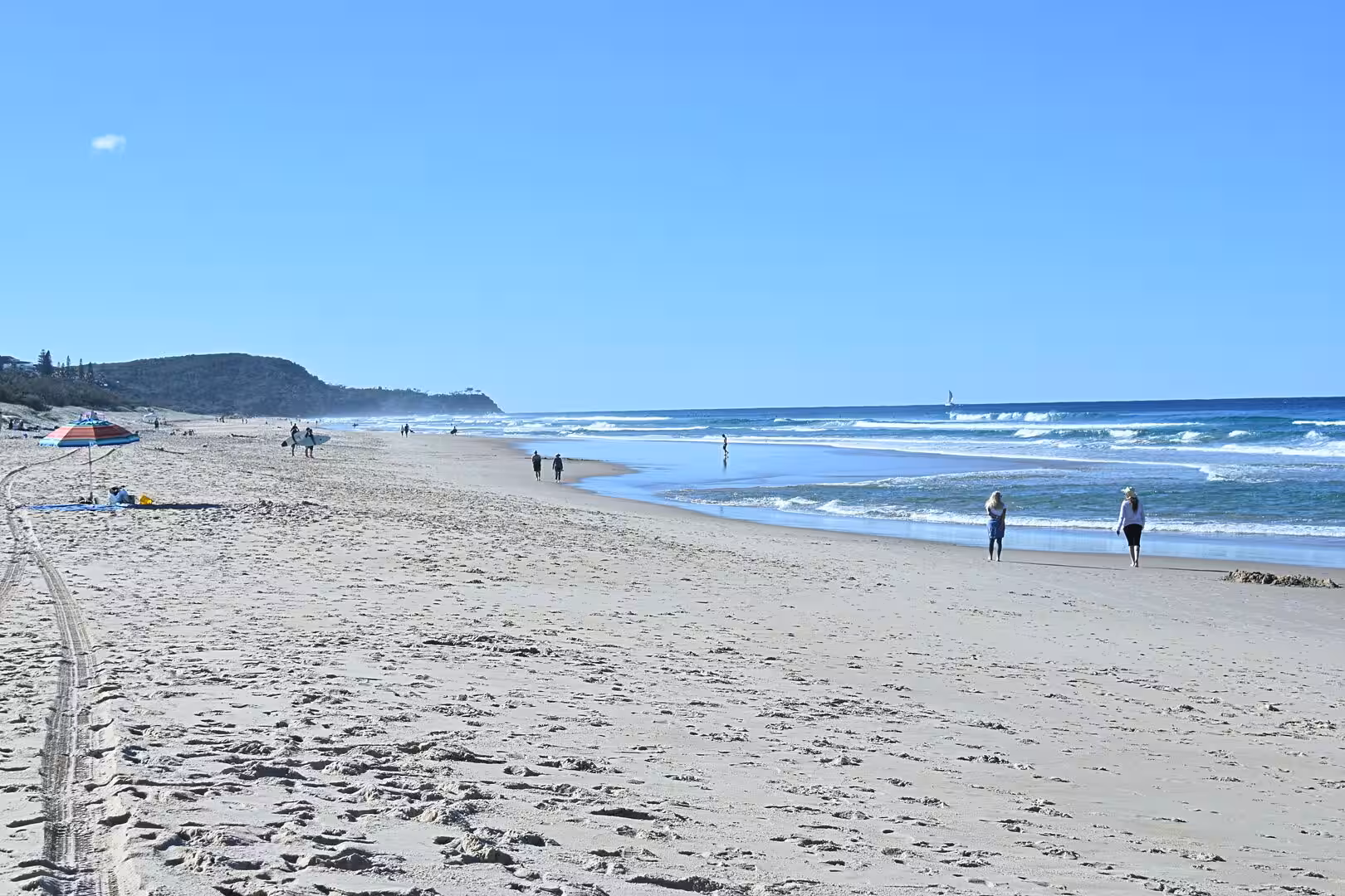 A wide sandy beach under clear blue sky with gentle surf and a headland in the distance.