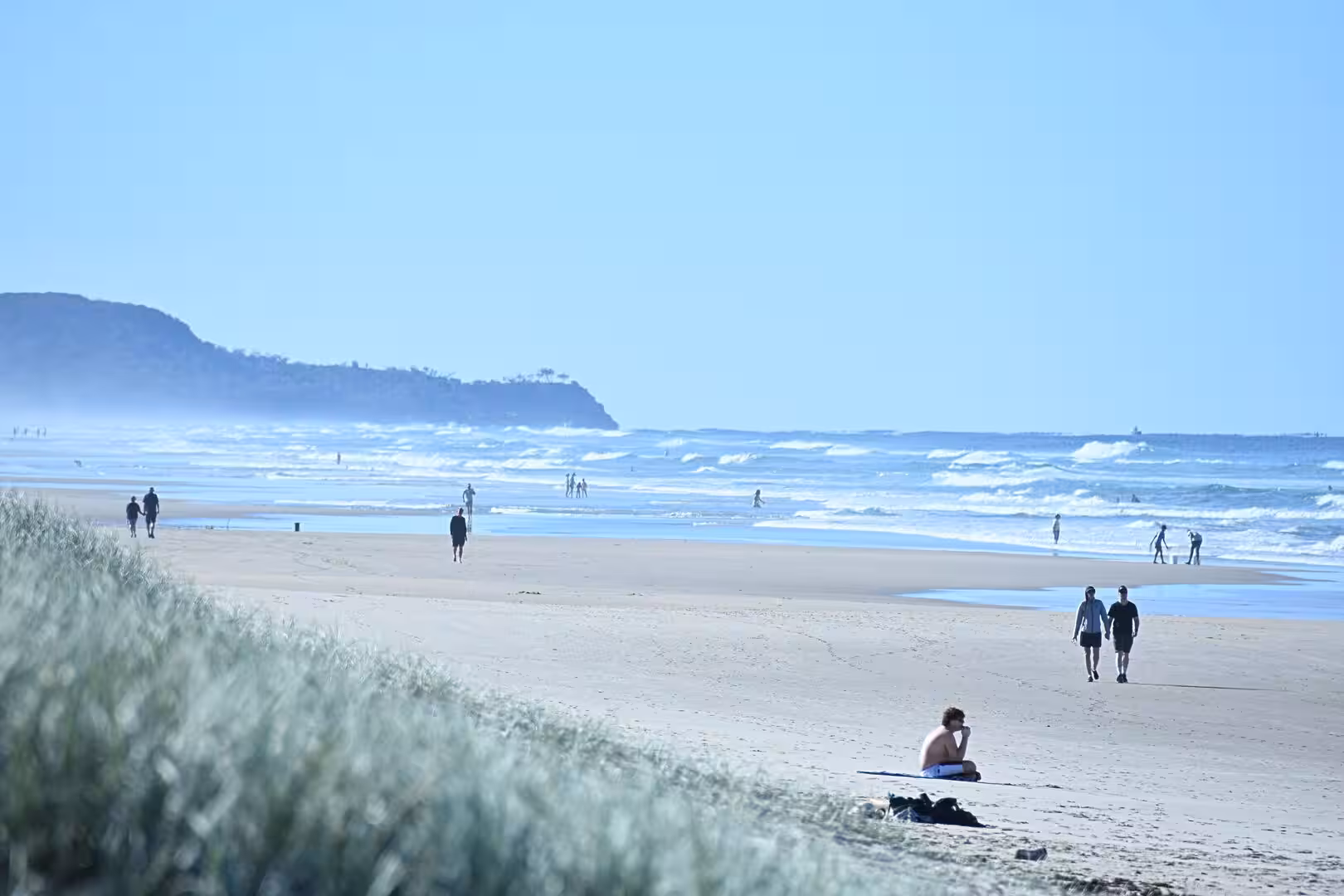 People walk and sit on a wide beach with surf and a distant headland, dune grass in the foreground.
