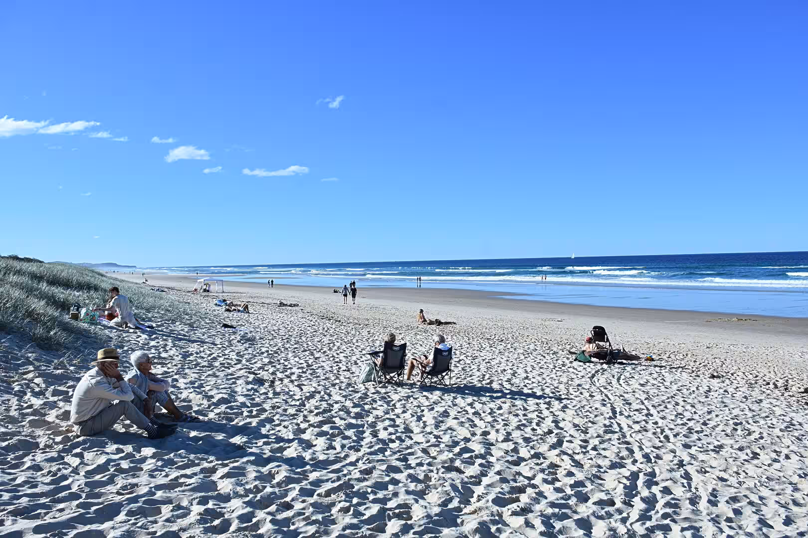 People sit and relax on a broad sandy beach under a blue sky with scattered clouds.