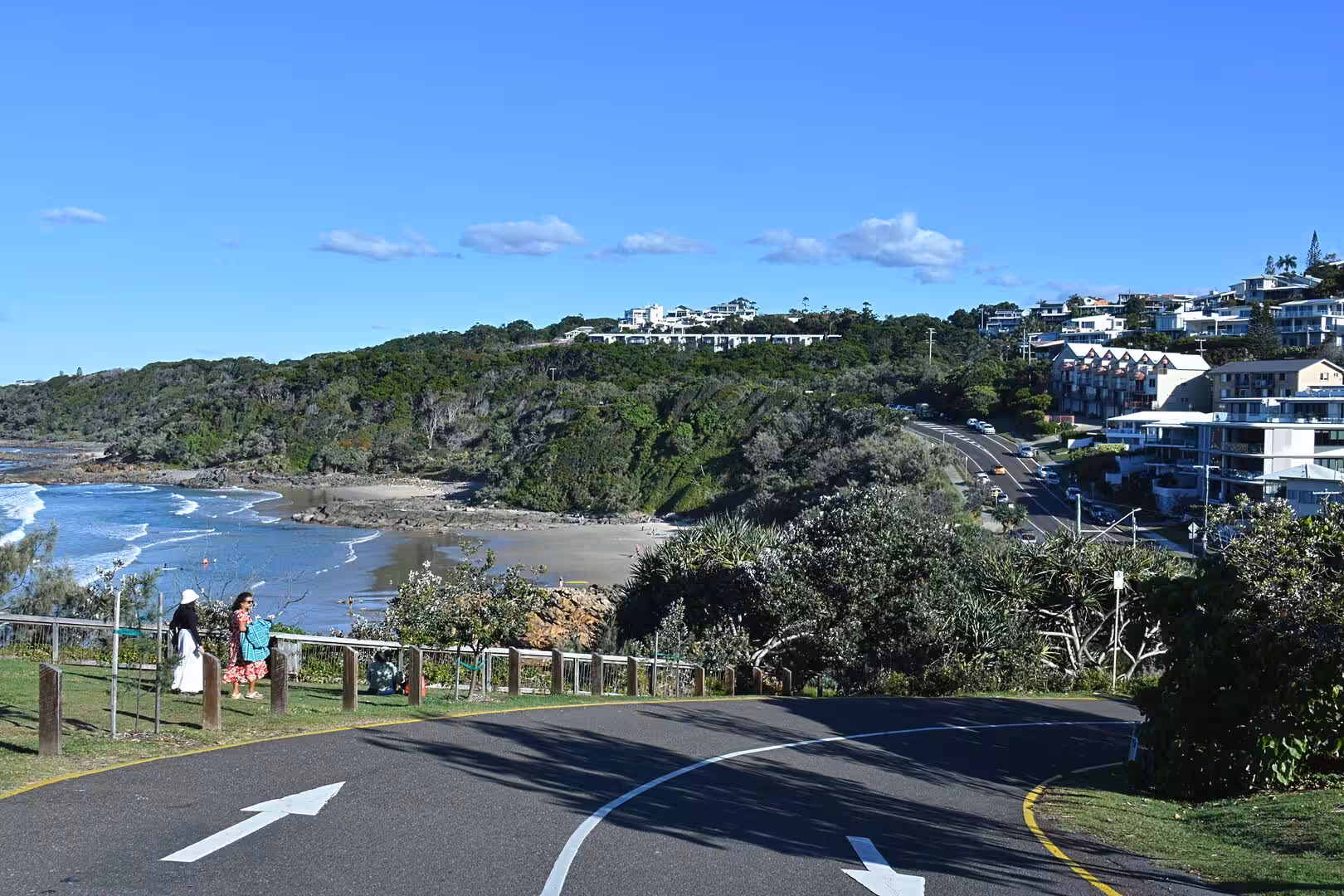 Visitors watch a small cove from a fence railing as a curved road passes below.