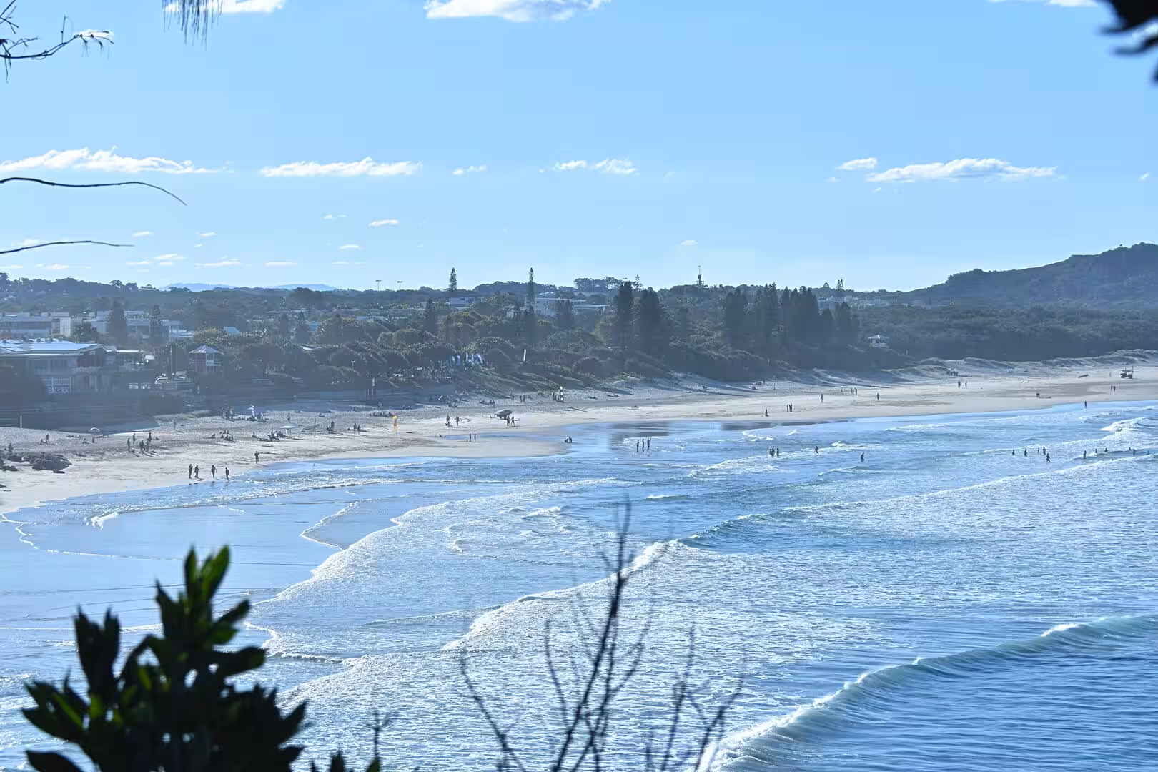 Many people walk a long sandy beach while waves roll in and a tower stands on the far hill.