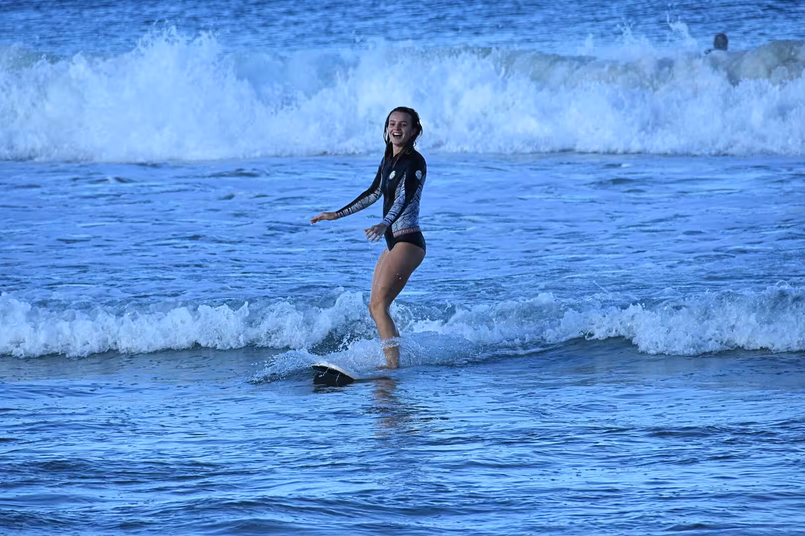 A smiling woman in a black wetsuit stands balanced on a surfboard riding a wave.