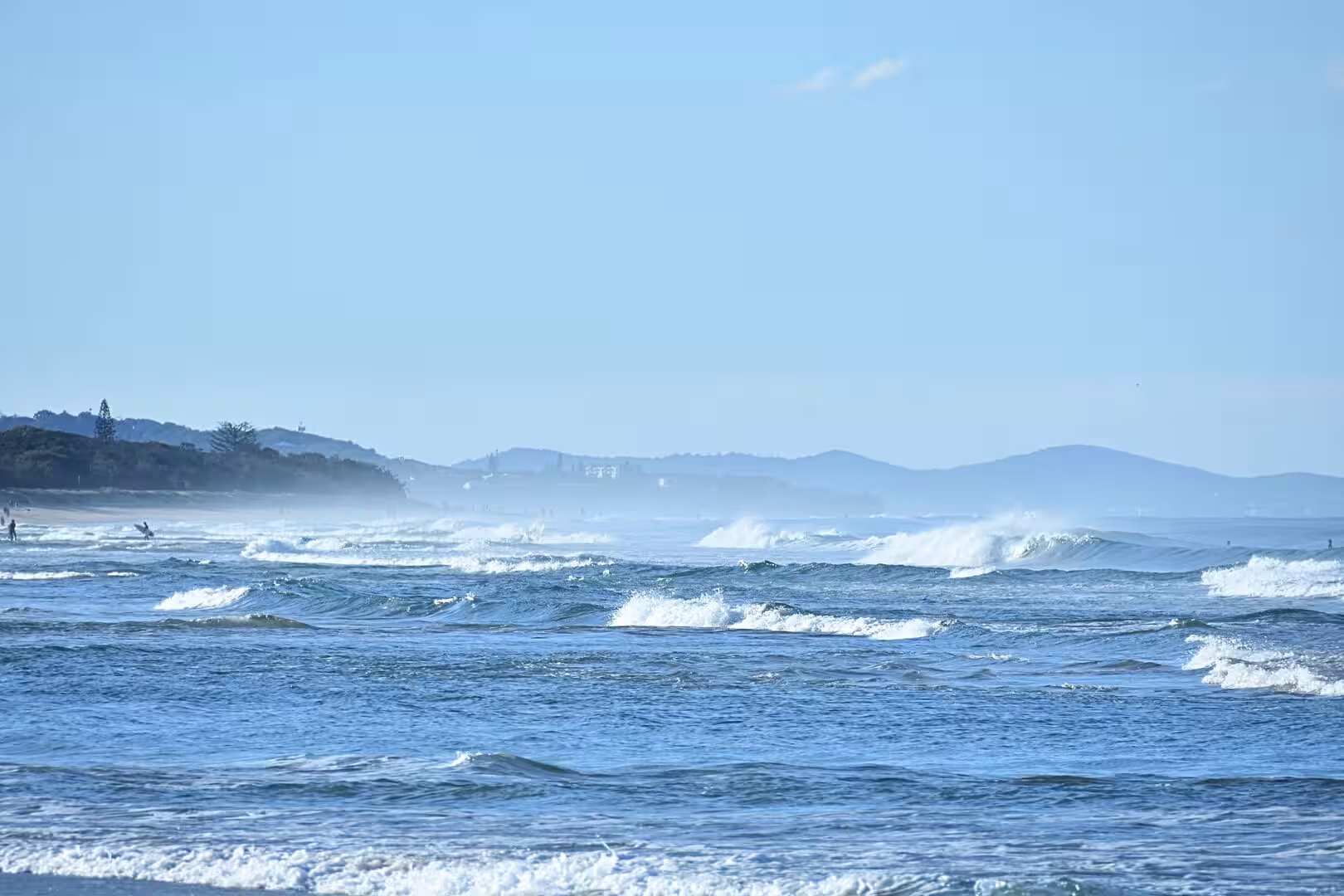 Lines of white surf break across a beach beneath a hazy blue sky.