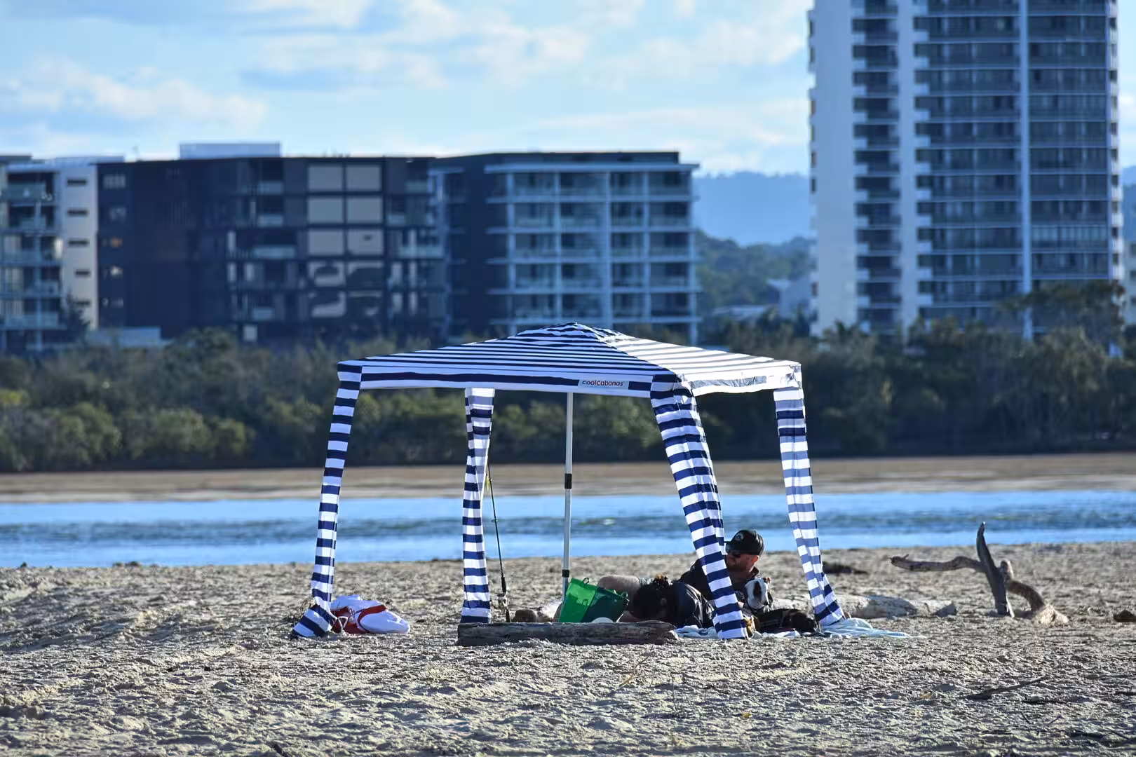 A blue-and-white striped beach tent shades people relaxing on the sand.