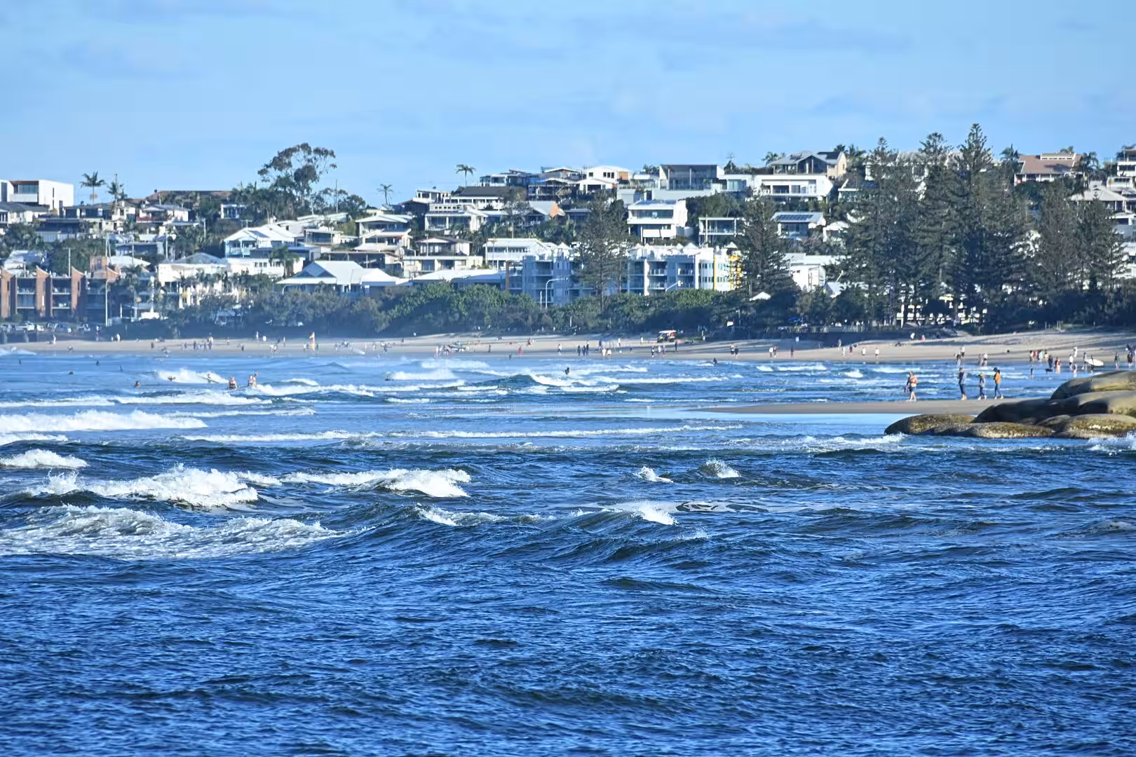 Dark blue water leads to a beach and a hillside suburb of houses among trees.