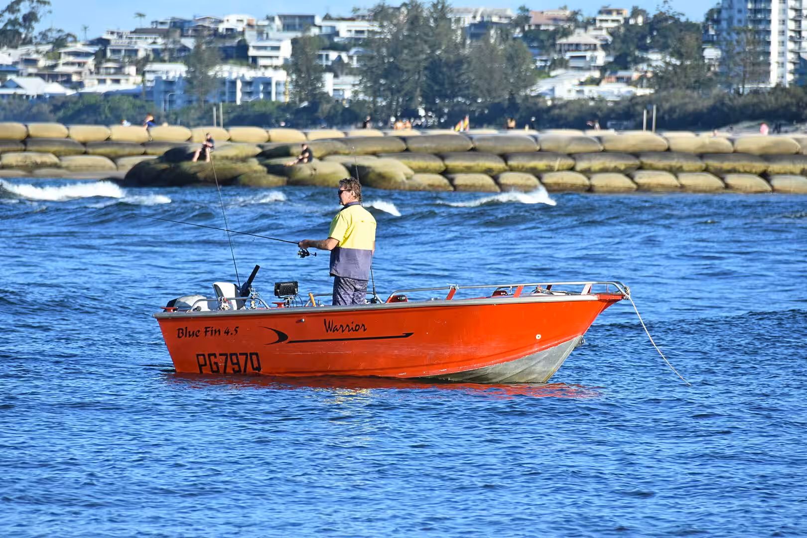 A man in a yellow shirt stands fishing from a bright red motorboat on blue water.