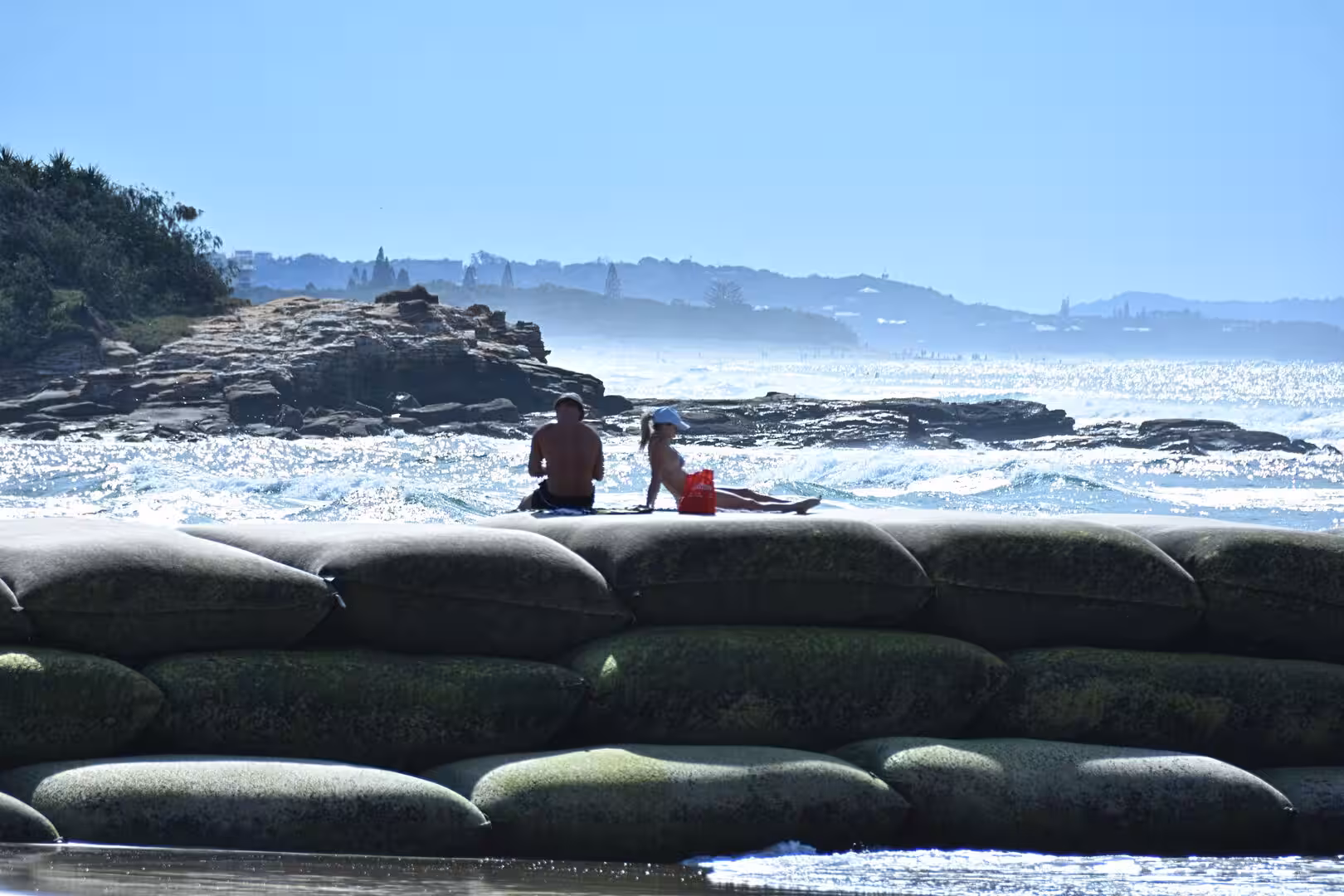 A man and a child in swimwear rest on the shore beside a wall of weathered green sandbags facing the surf.