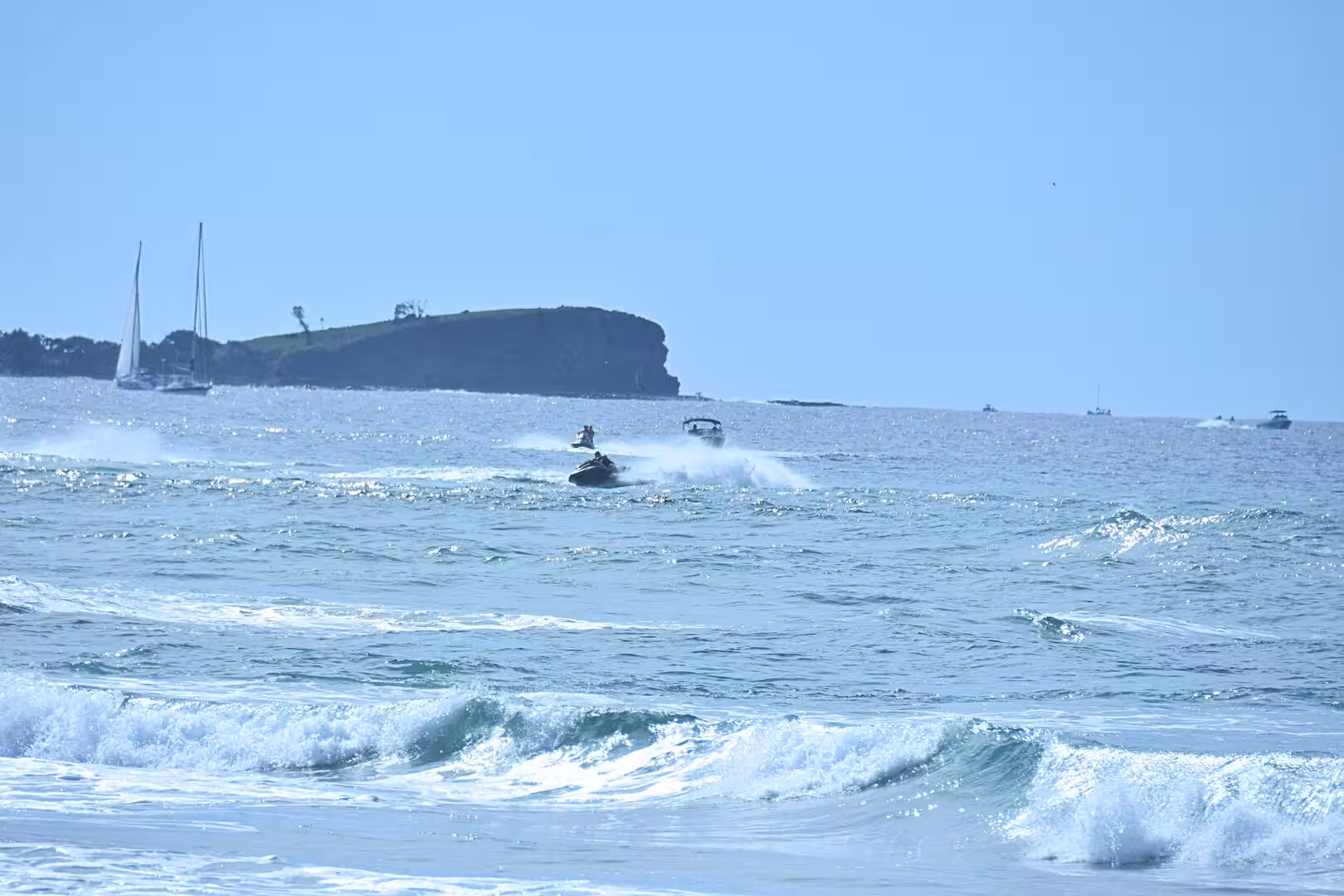 Small watercraft race across white-capped surf with a sailboat and a green headland in the hazy distance.