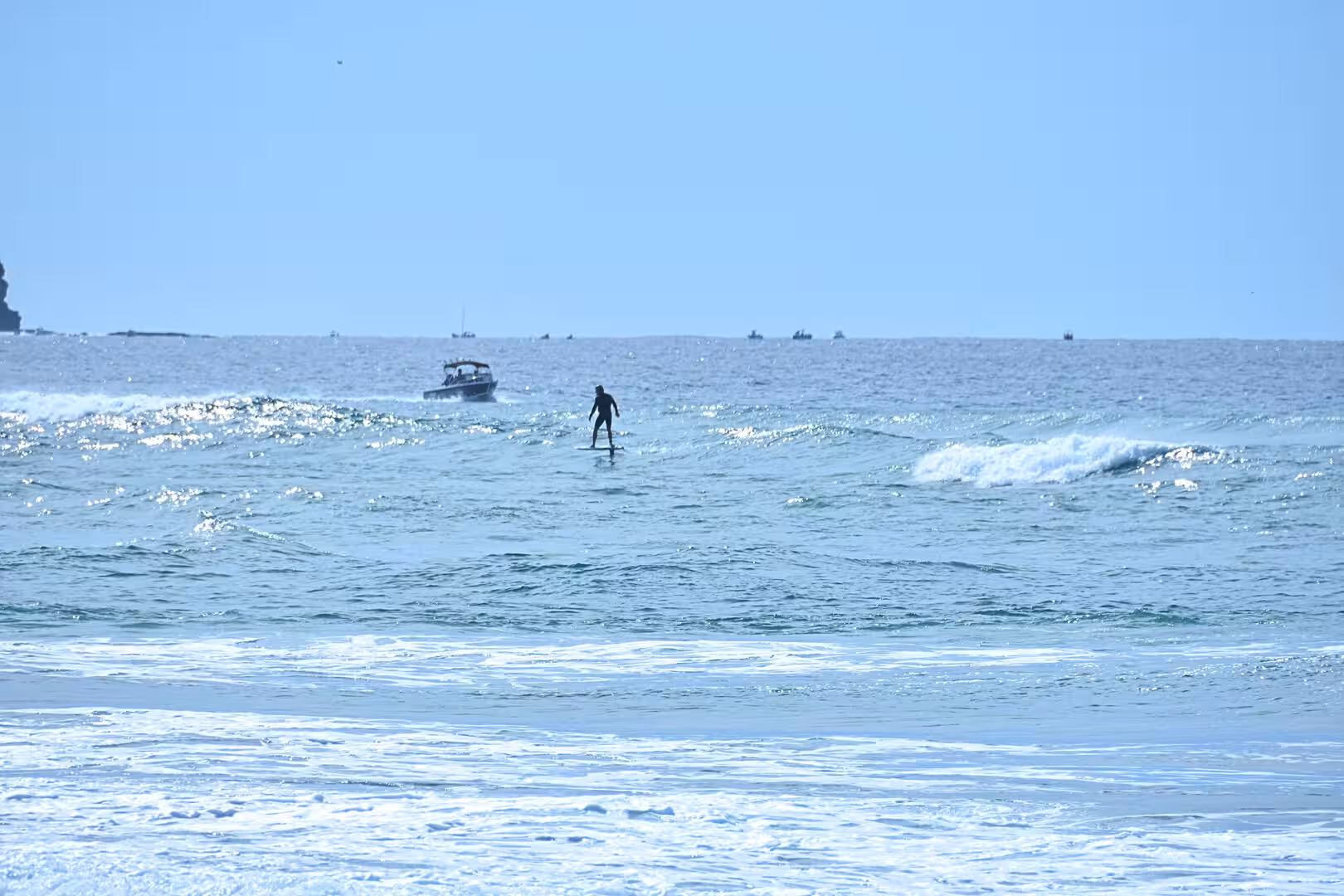 One person stands upright on a paddleboard while a motorboat and distant boats bob beyond the waves.