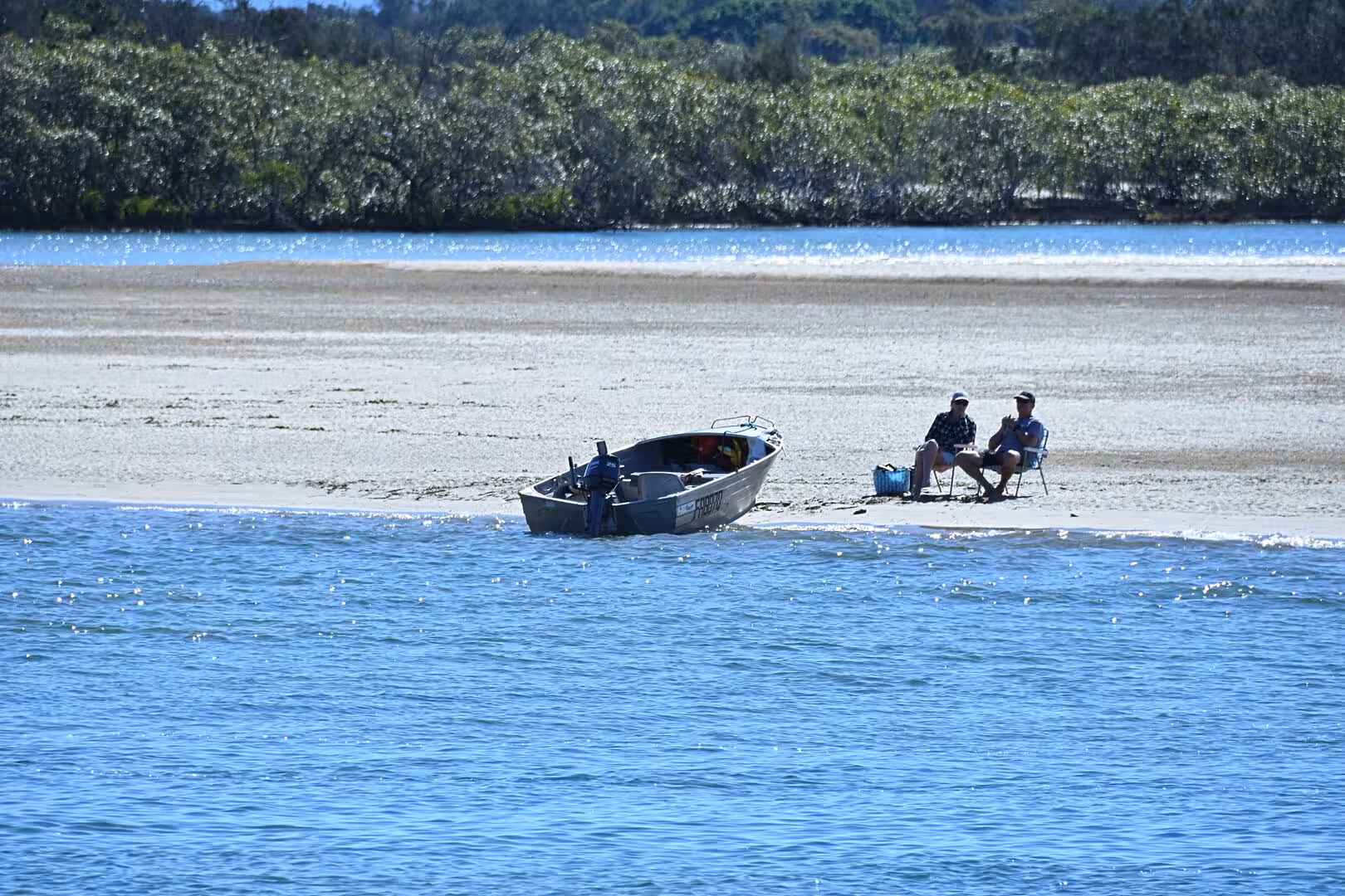 Two people sit in folding chairs on a low sandbank next to a small boat, with calm water and lush trees around.