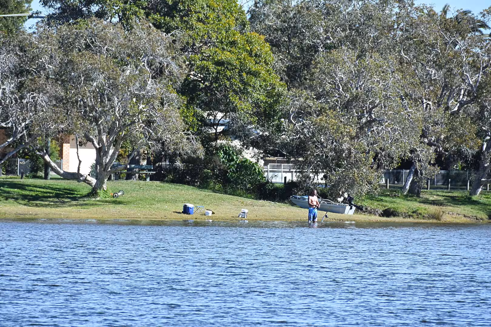 A solo paddler stands in shallow water near a grassy shore with a canoe and gear resting on the bank.