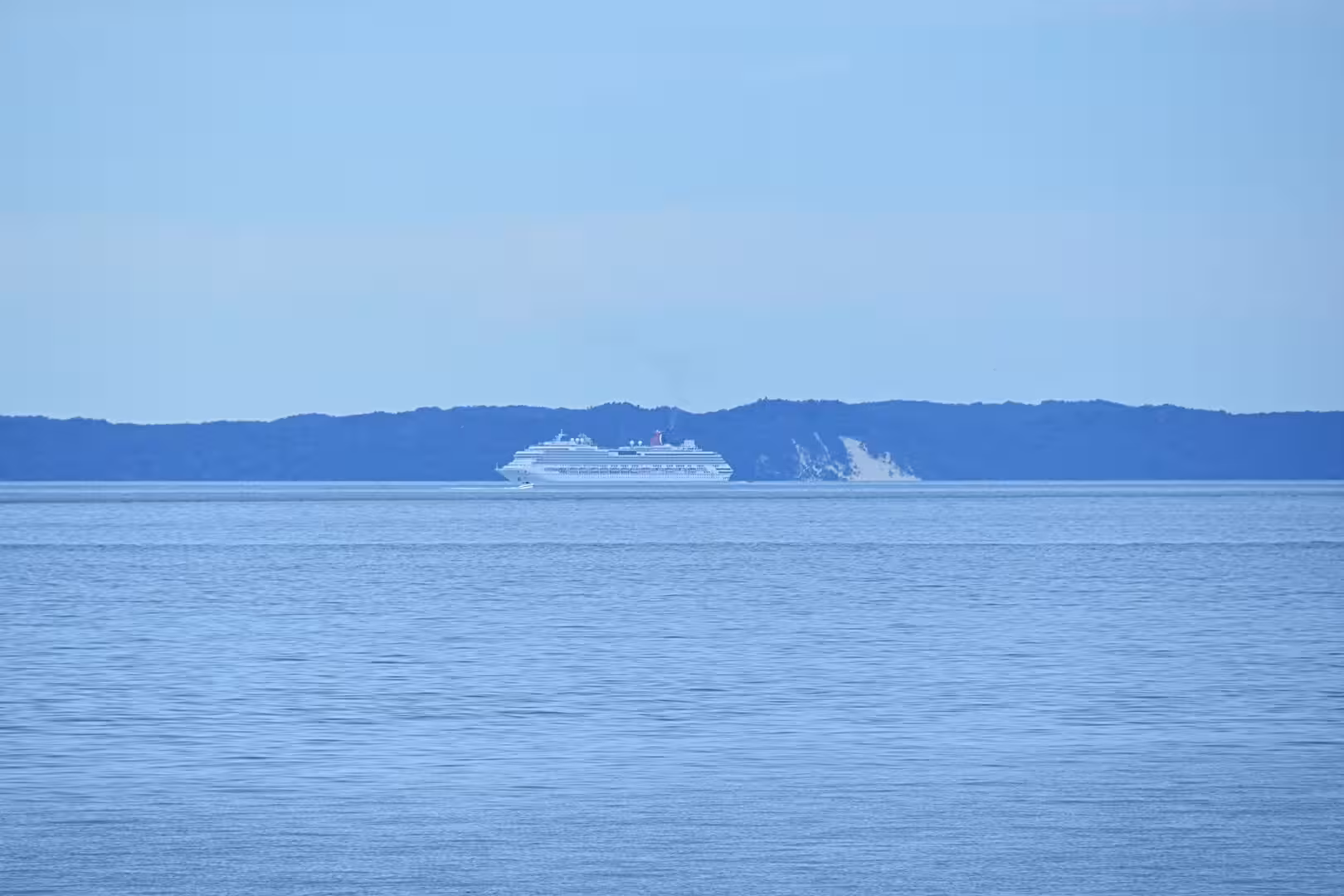A large white cruise liner crosses calm blue water while a dark forested cape rises on the horizon.