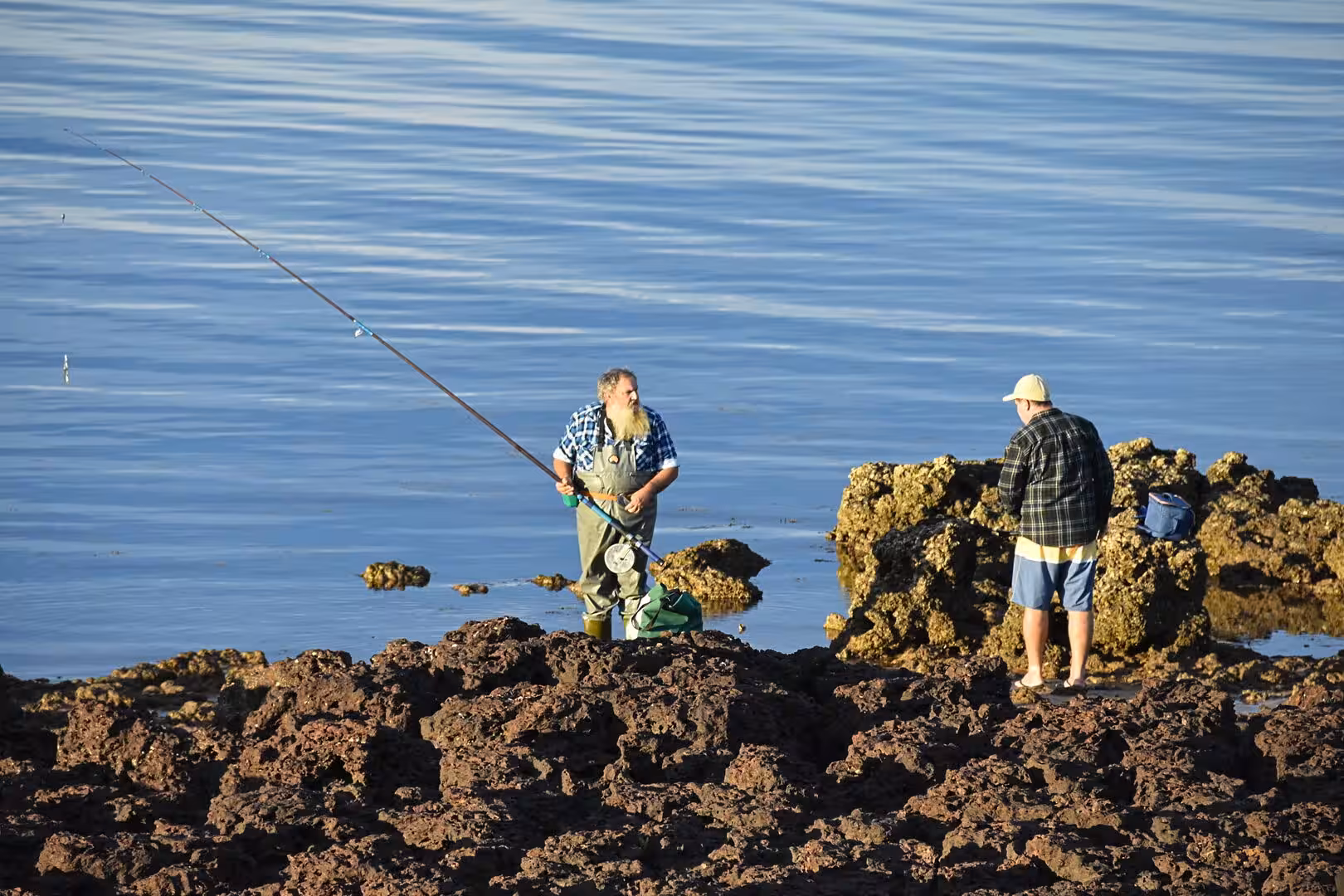 Two men fish from a rocky shore on a calm blue sea.
