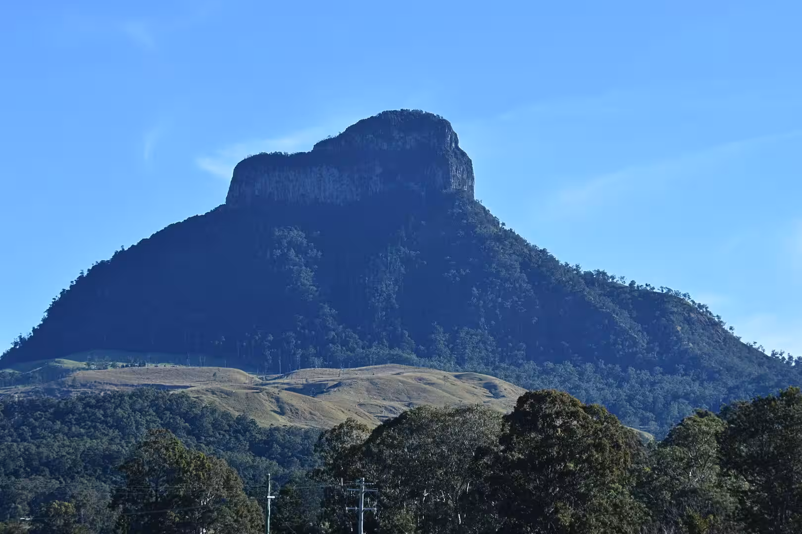 A sheer forested mountain rises above rolling green hills against a blue sky.