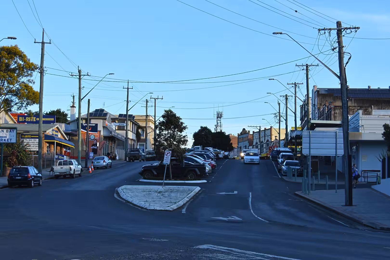 A quiet suburban street lined with shops and parked cars at dusk.