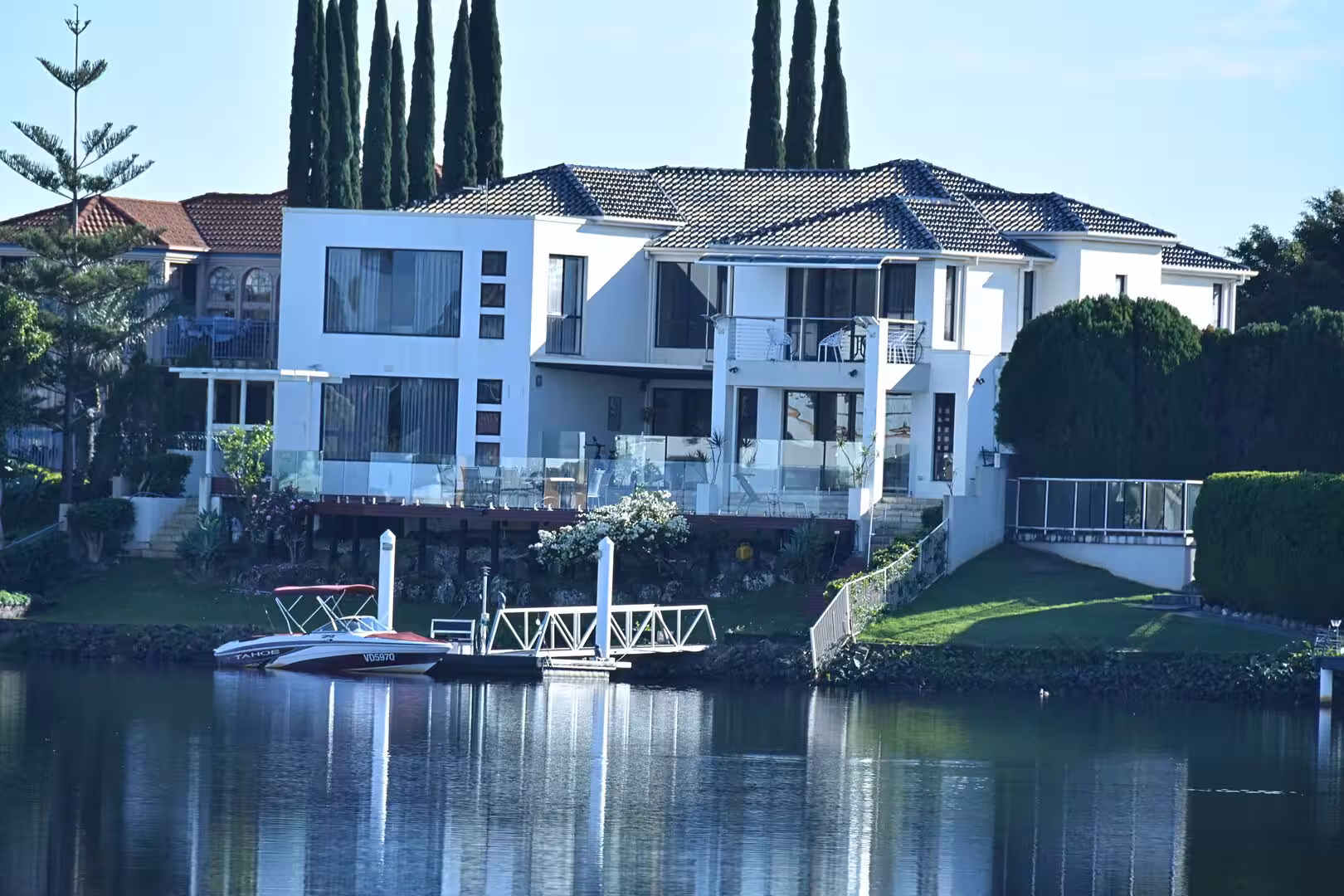 A tall white waterfront home stands behind cypress trees and a boat dock.