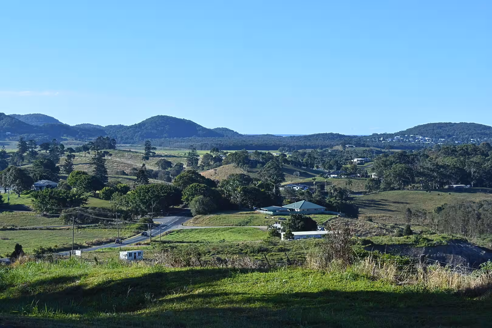 A winding road cuts through green paddocks beneath distant blue hills.
