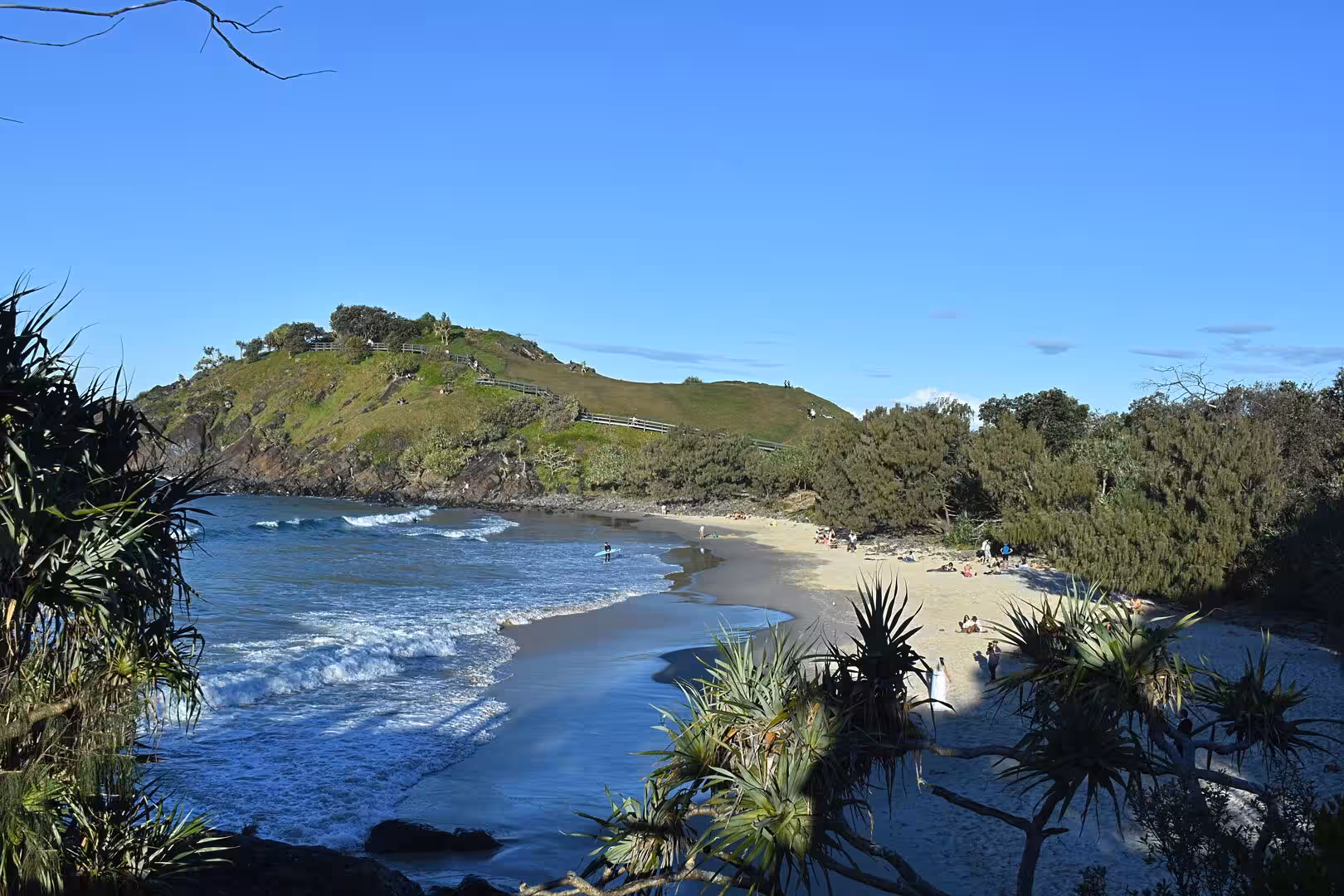 A green coastal hill with a walking track rises above a sandy beach.