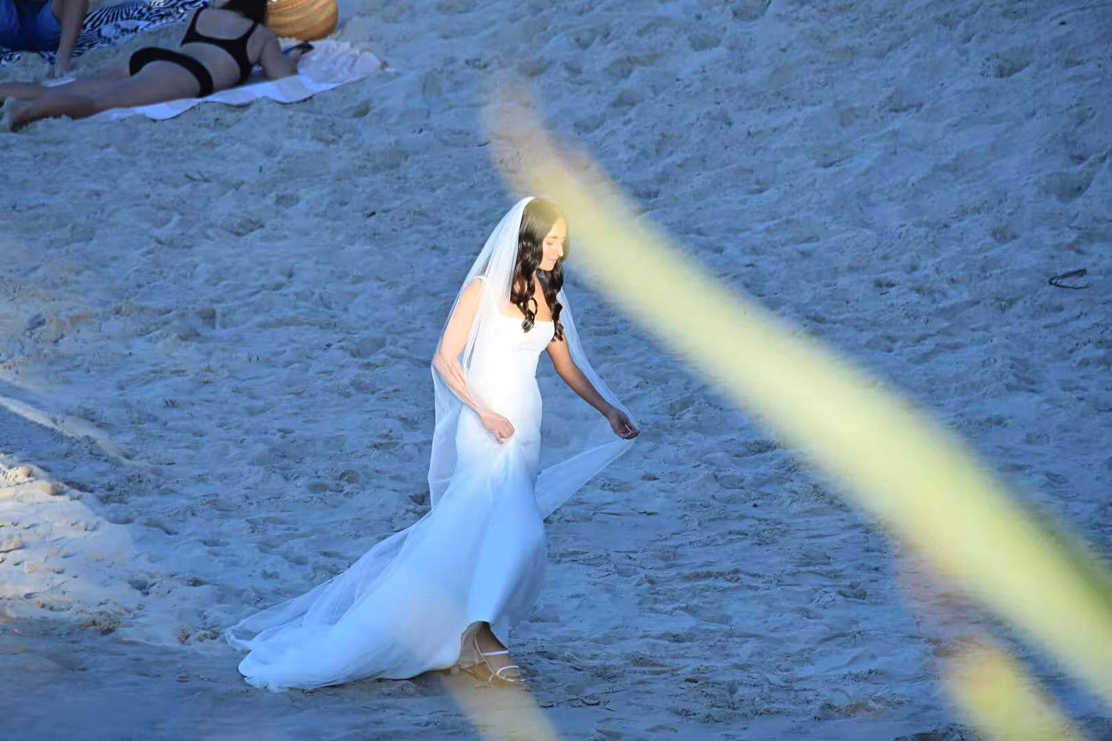 A bride in a white gown steps along the sand by the sea.