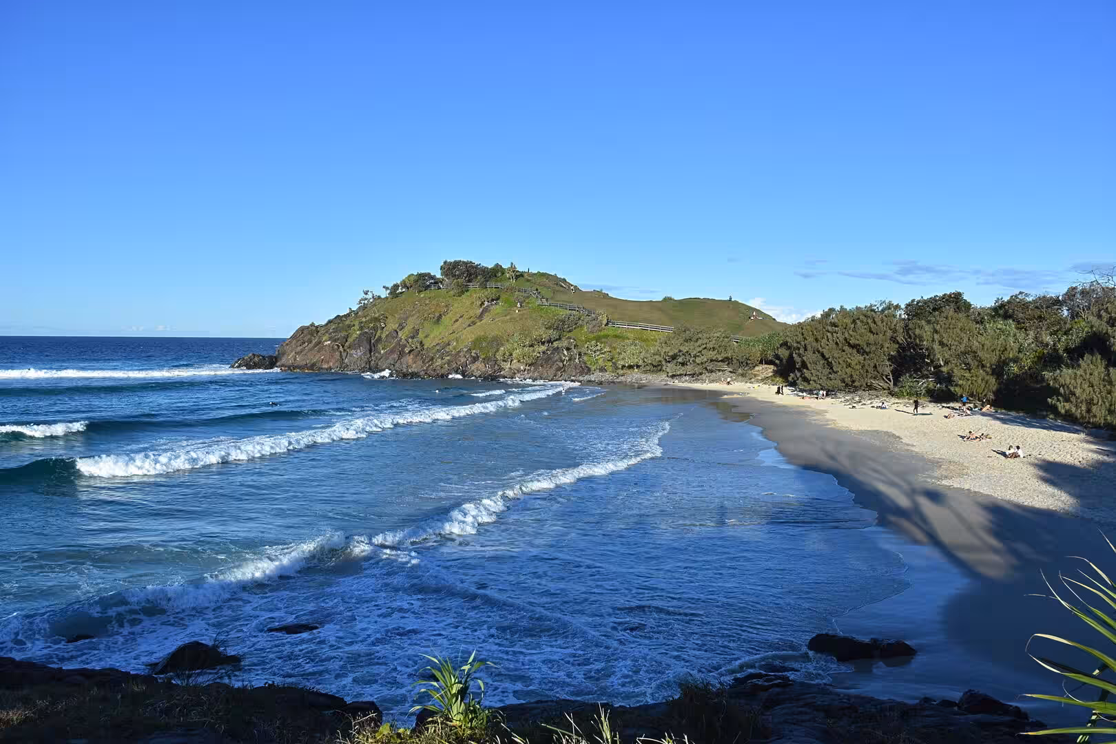 Waves roll onto a sandy beach beneath a green, rocky headland.