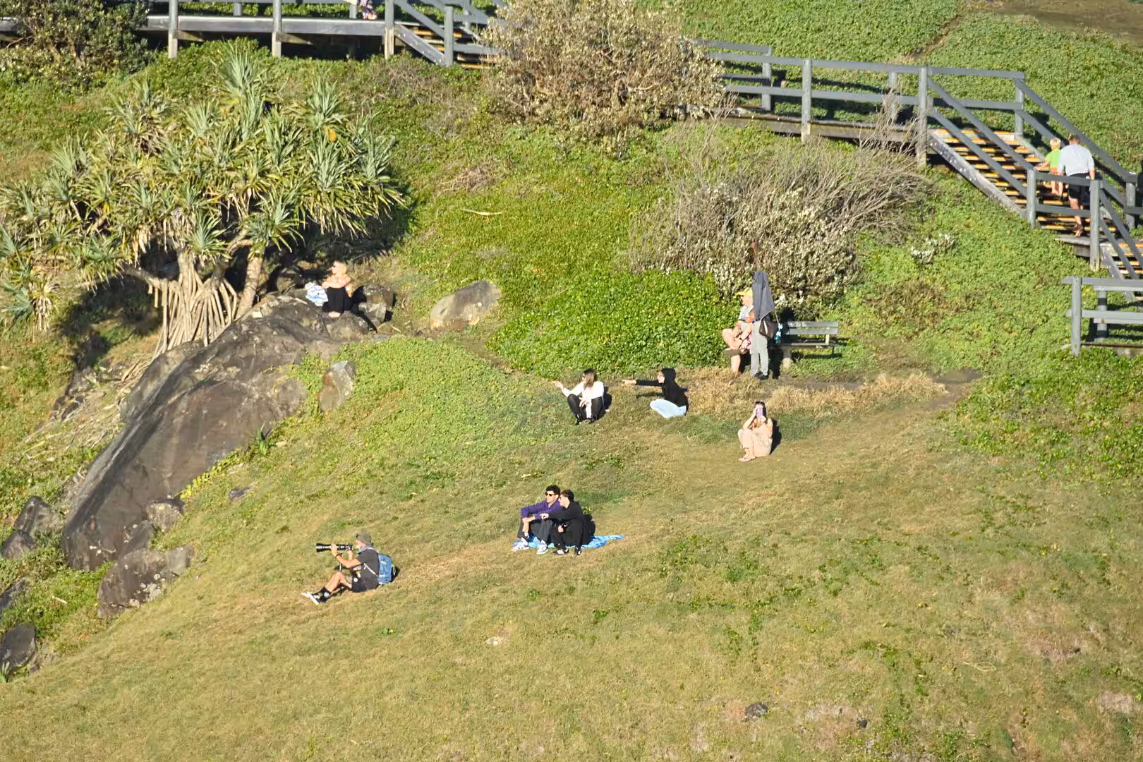 Visitors sit and stroll on a rocky grassy slope with wooden walkways.