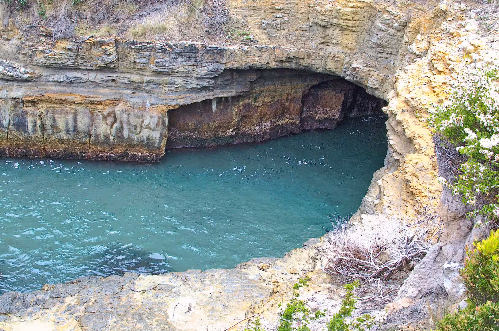 Layered sandstone cliffs form a dark cave opening over a clear turquoise rock pool.