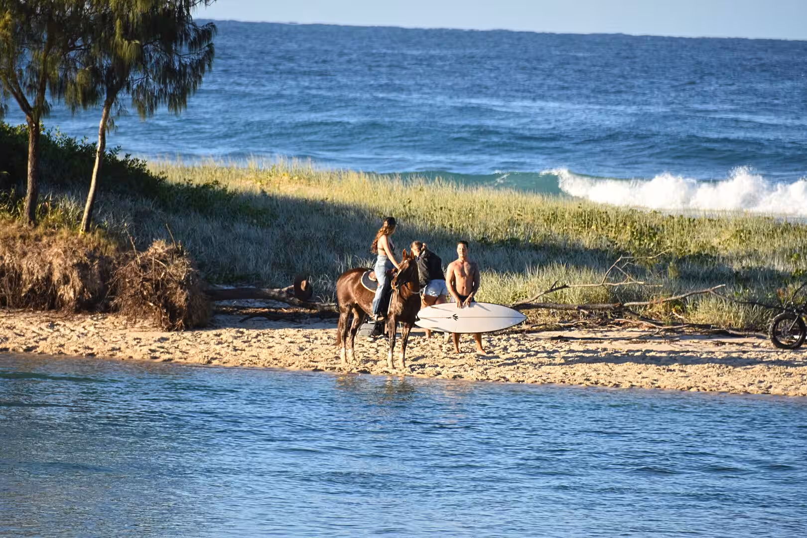 A bareback rider on a horse stands with surfers holding a board on the sand.
