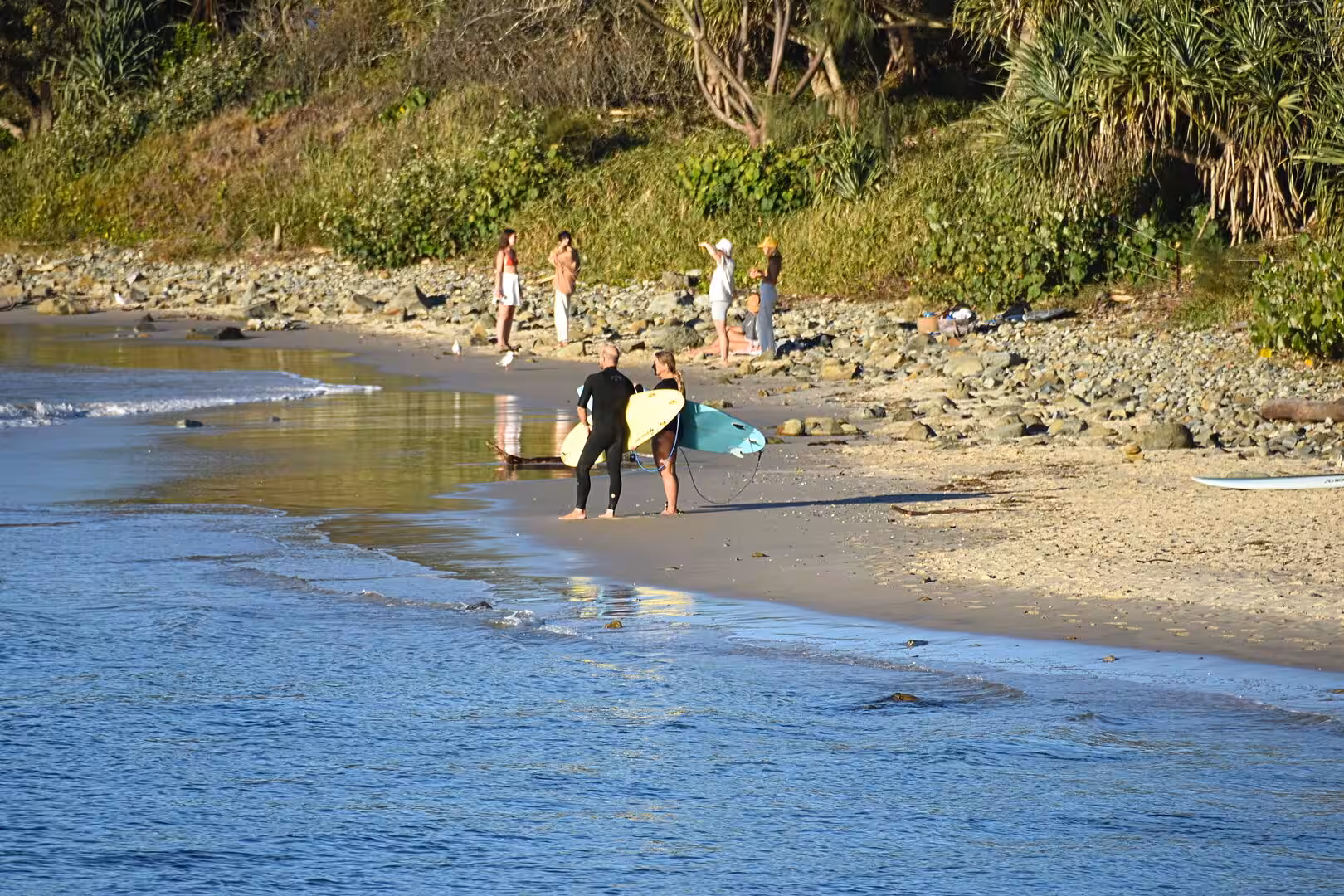 Two surfers carry their boards at the water's edge while others relax on the beach.