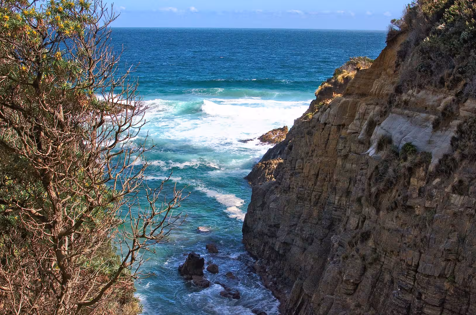 Waves crash against a steep coastal cliff beneath a deep blue sky.