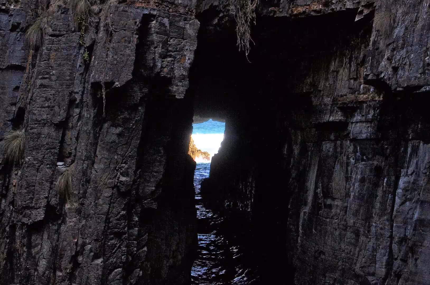 Bright sea and sky are framed through a narrow gap between dark cliff walls.