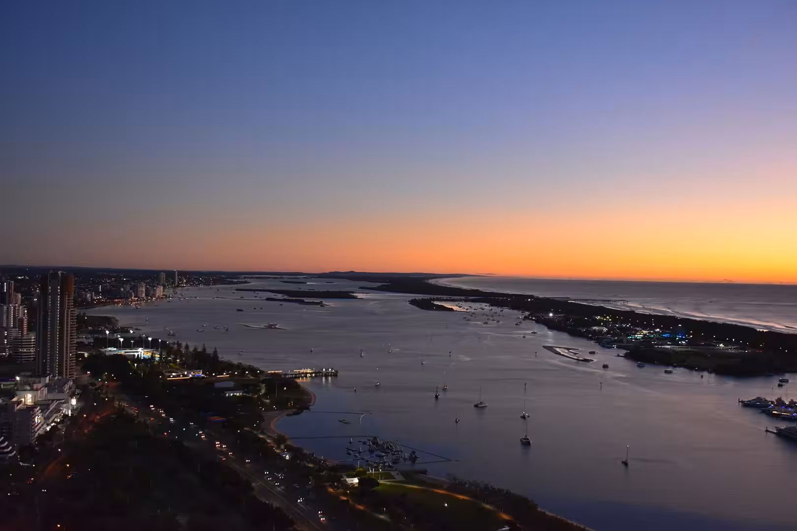 From above, boats and a skyline are silhouetted against a glowing harbour.