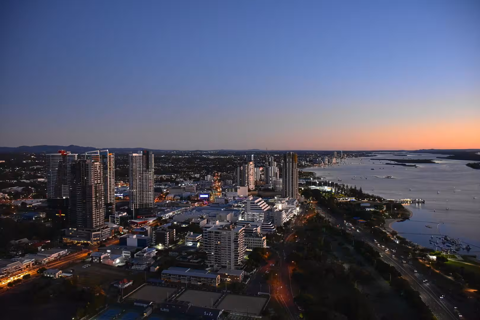 Towering skyscrapers stand lit while a calm bay fades into warm evening light.