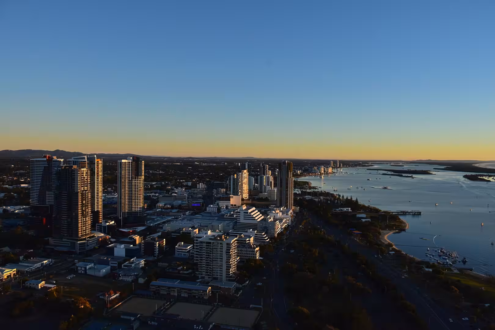 Tall modern skyscrapers rise beside a broad bay dotted with small boats at sunset.