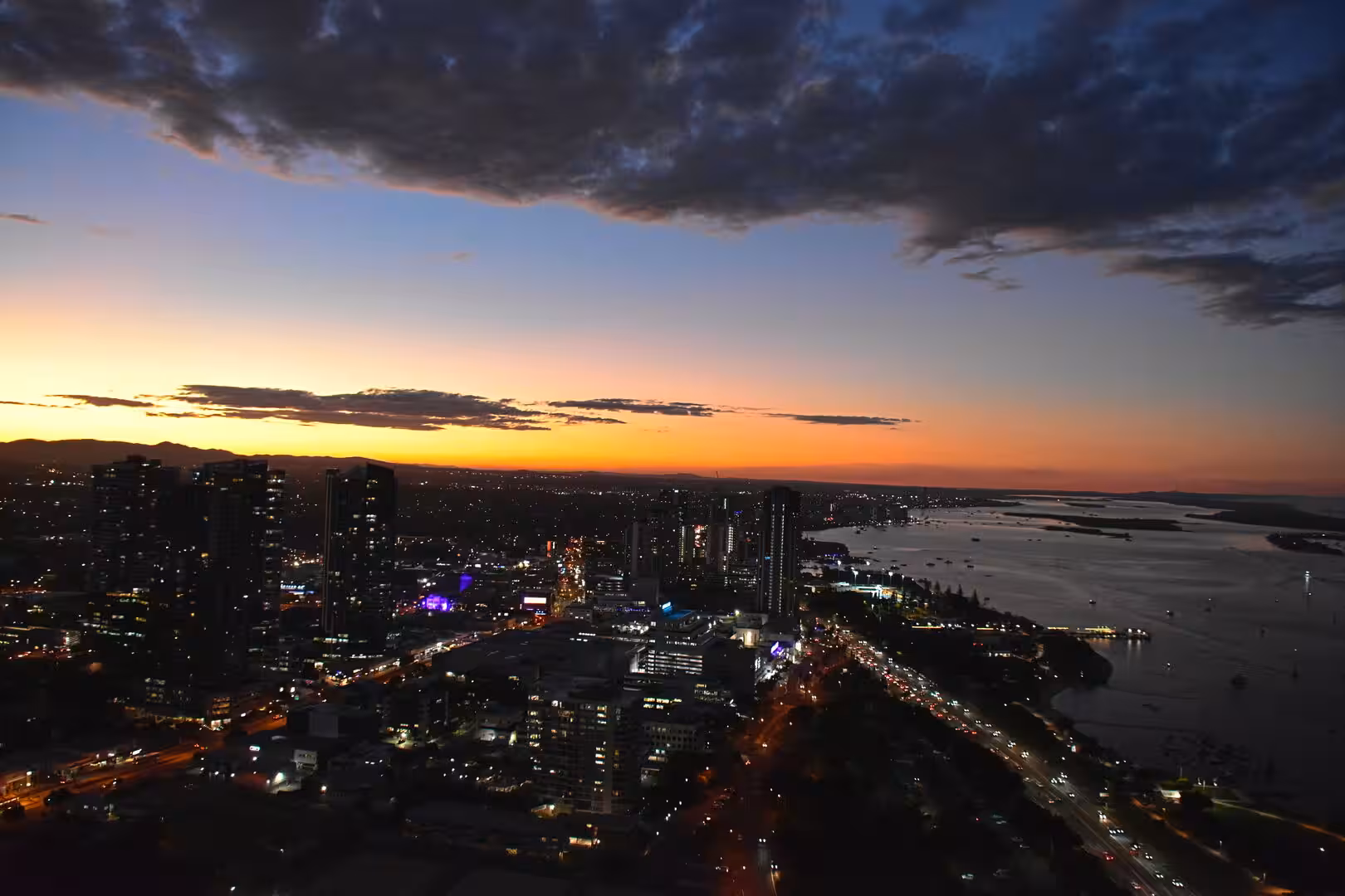 A long bridge spans the water as the city lights up under a fiery orange sunset.