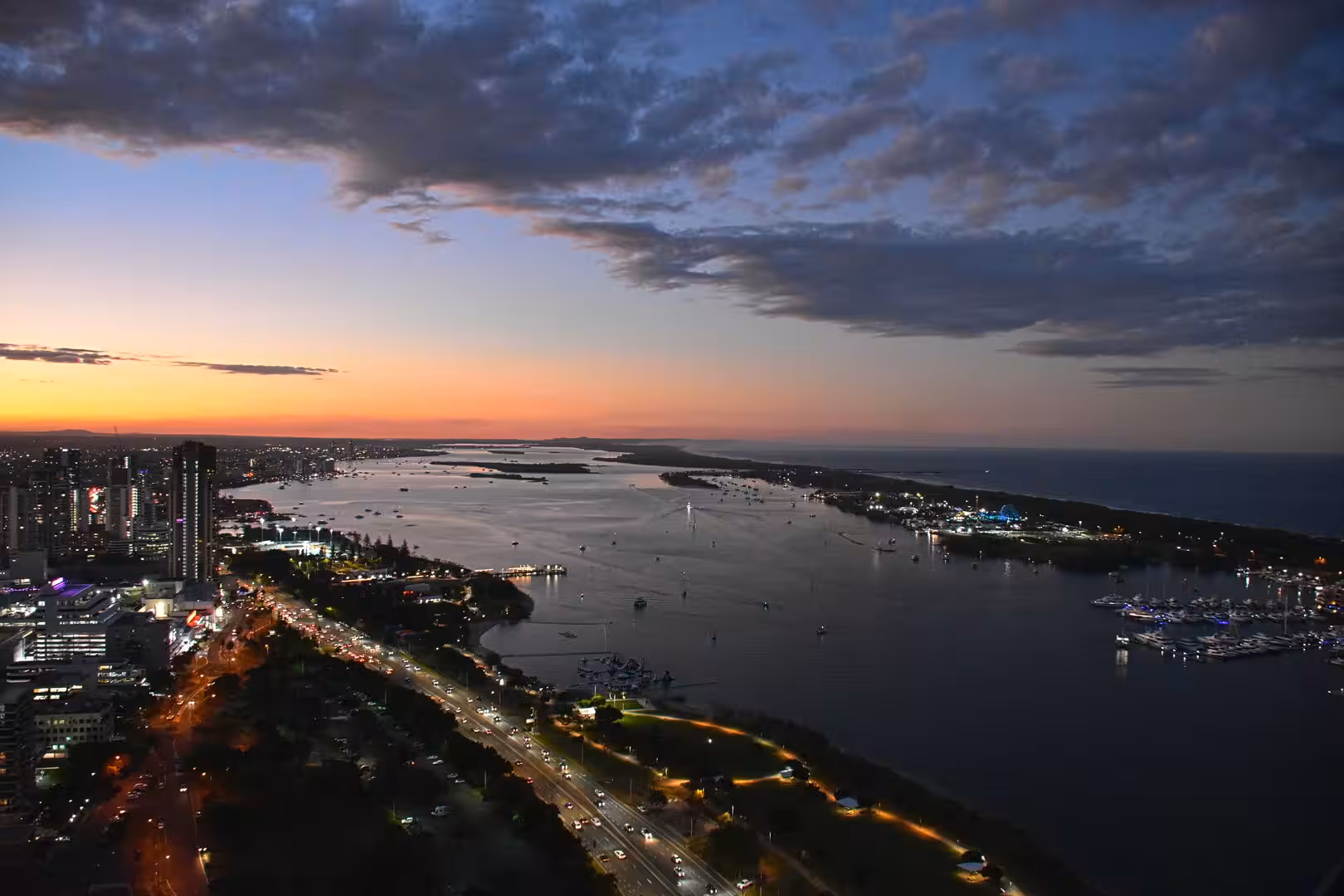 A twilight aerial view of a coastal harbor dotted with moored boats and glowing city lights.