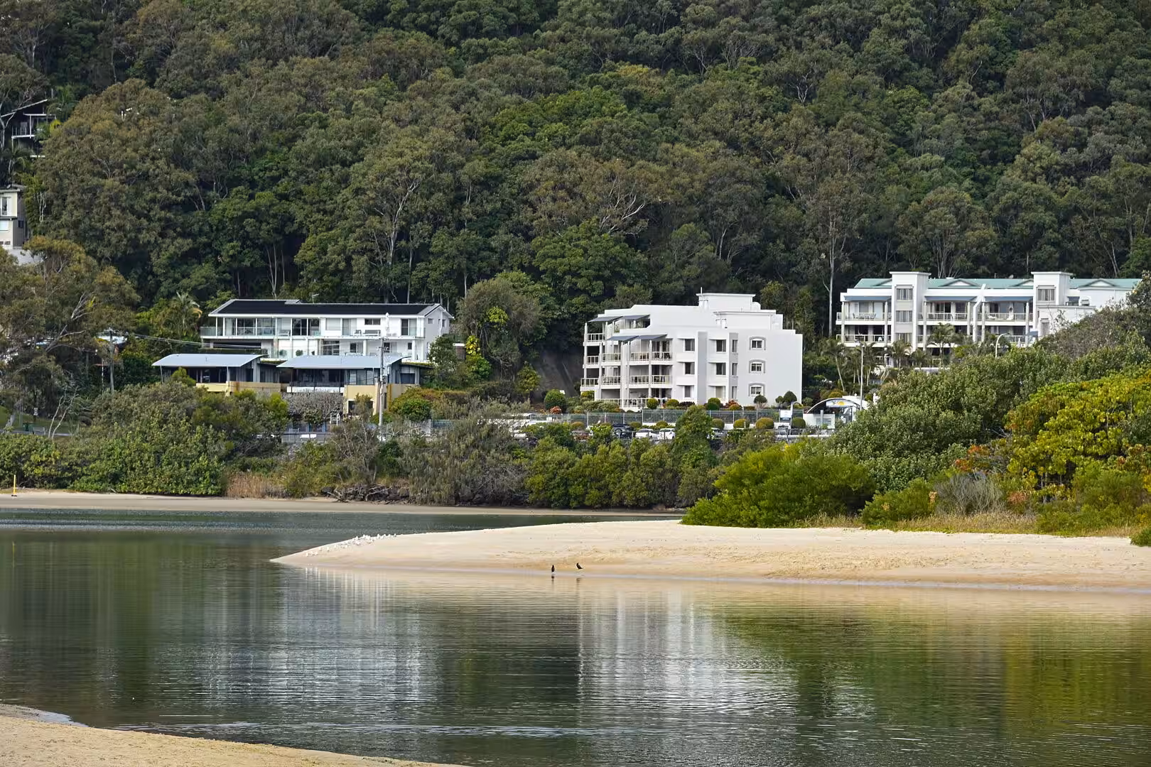 White coastal houses line the foot of a forested hill above a calm, mirror-still inlet.