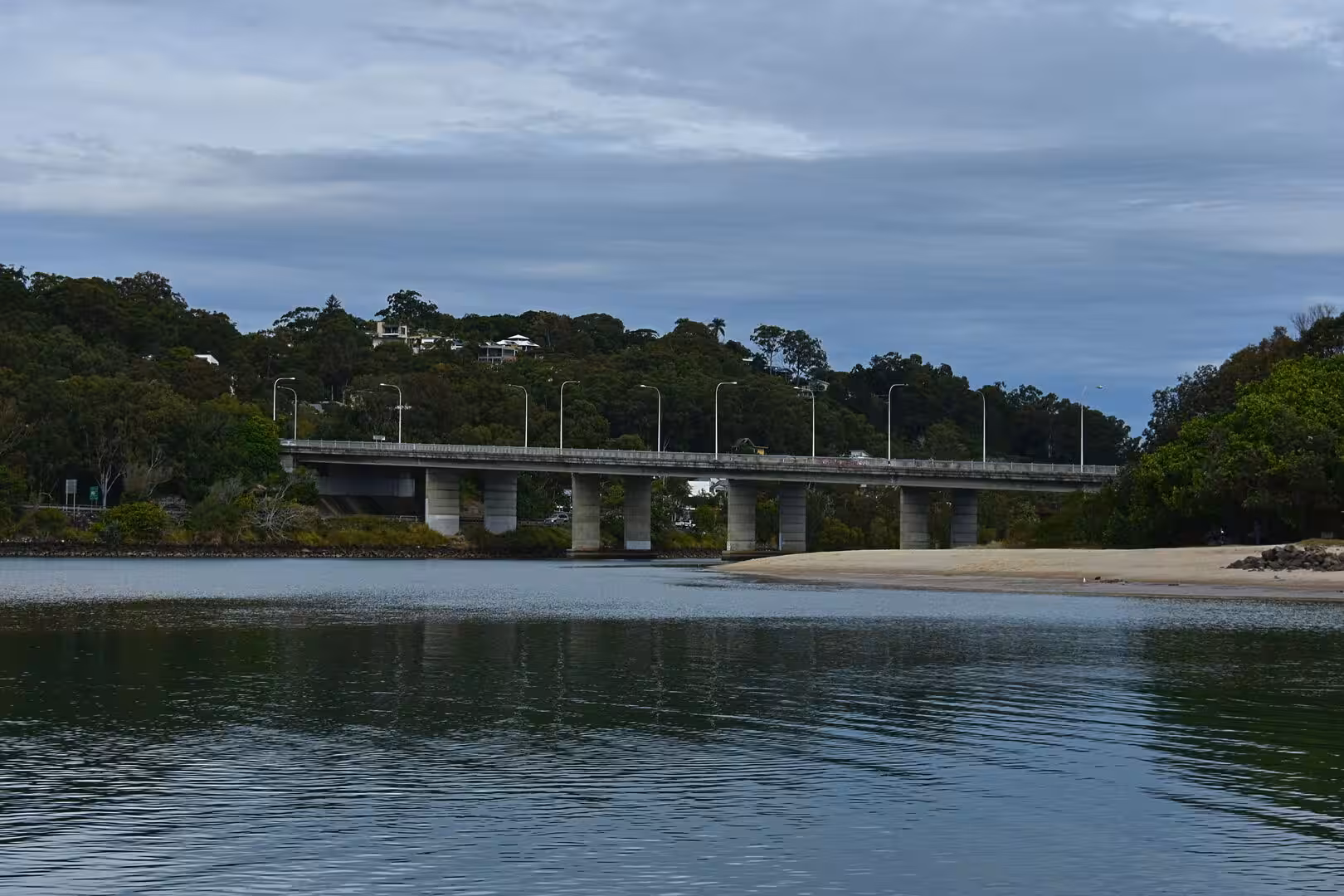 A concrete bridge with streetlights spans a calm inlet beneath forested hills and a cloudy sky.