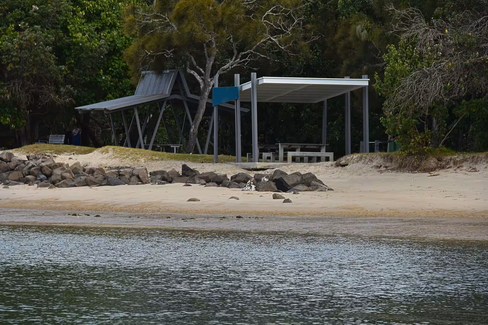 Covered picnic shelters stand on a sandy beach behind a rocky breakwater and calm water.