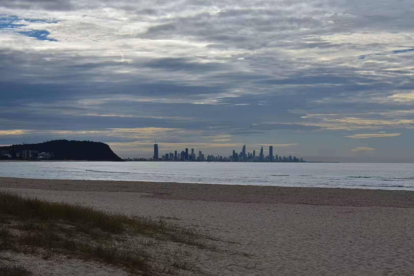 A sandy beach with dunes looks across calm water to a distant city skyline.