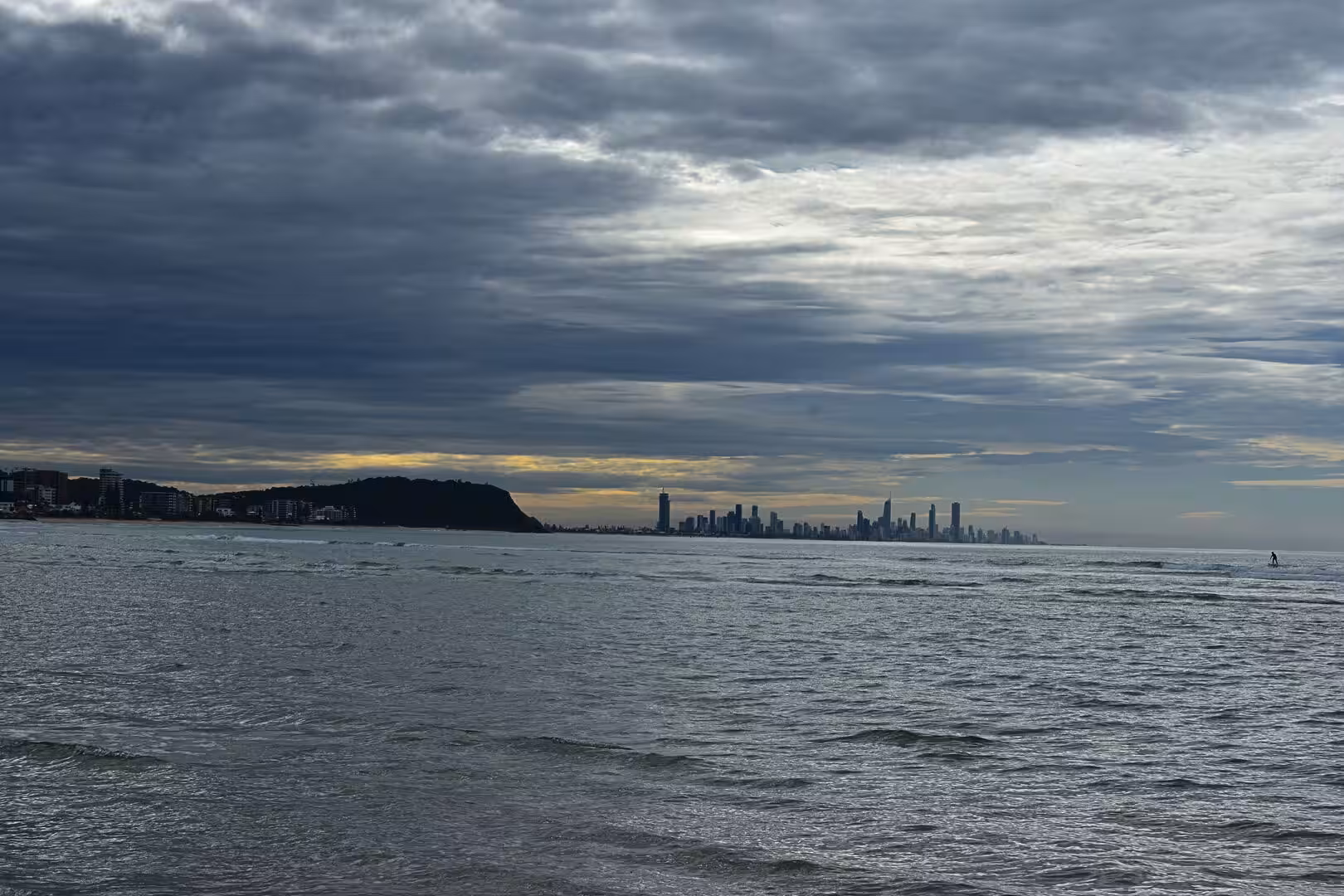 A choppy grey sea stretches toward a distant skyline with a single swimmer in the water.