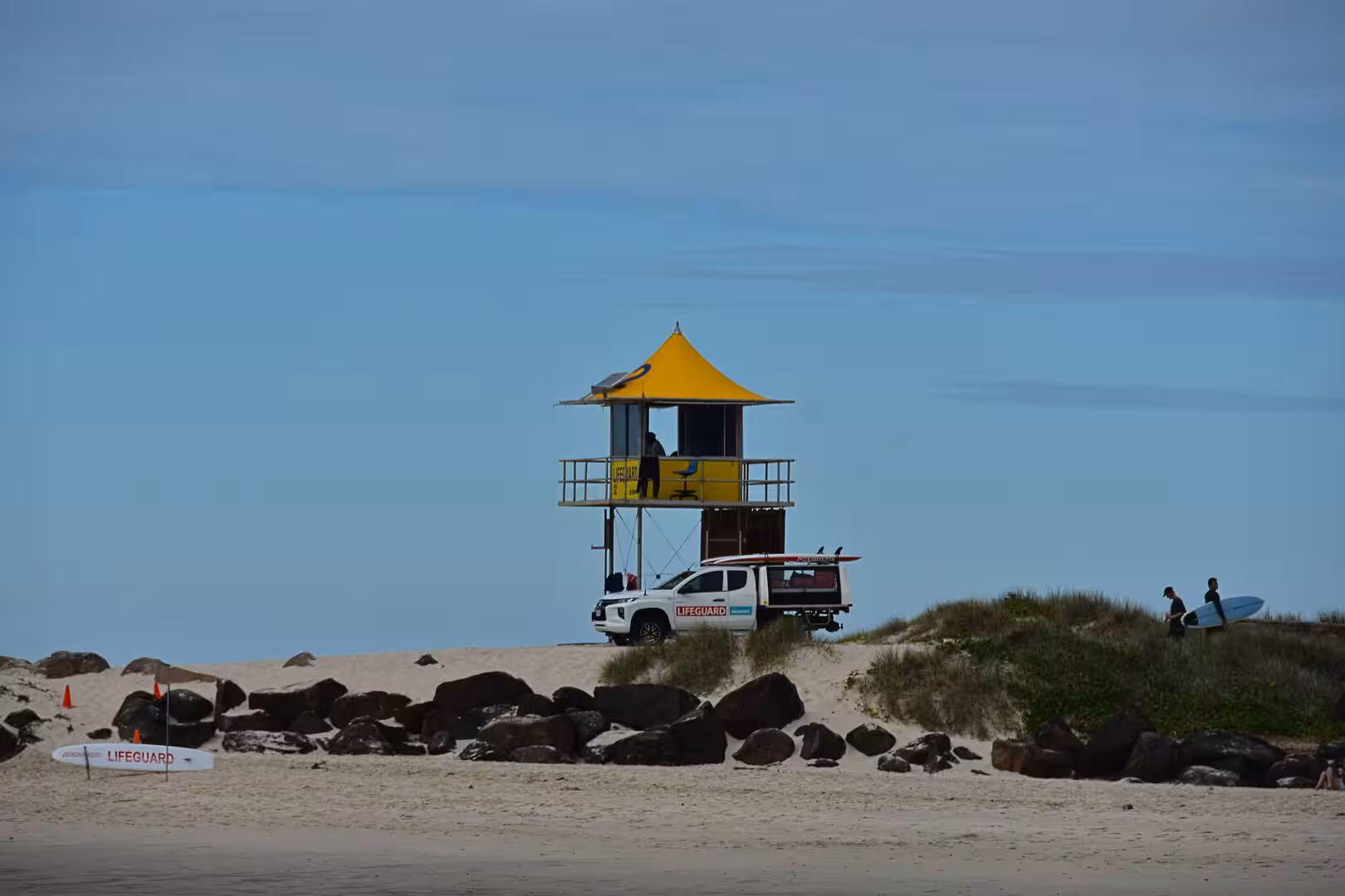 A yellow-roofed lifeguard tower stands on the beach with a white patrol vehicle and surfboards.
