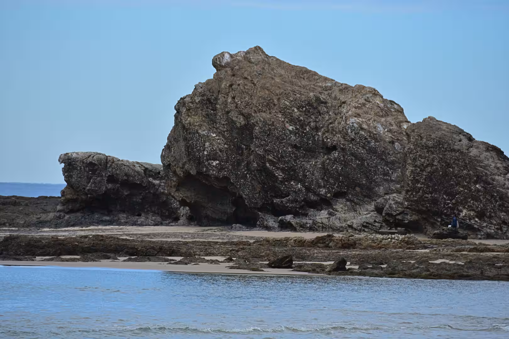 A large craggy rock rises from a seaweed-strewn beach beside calm blue water.