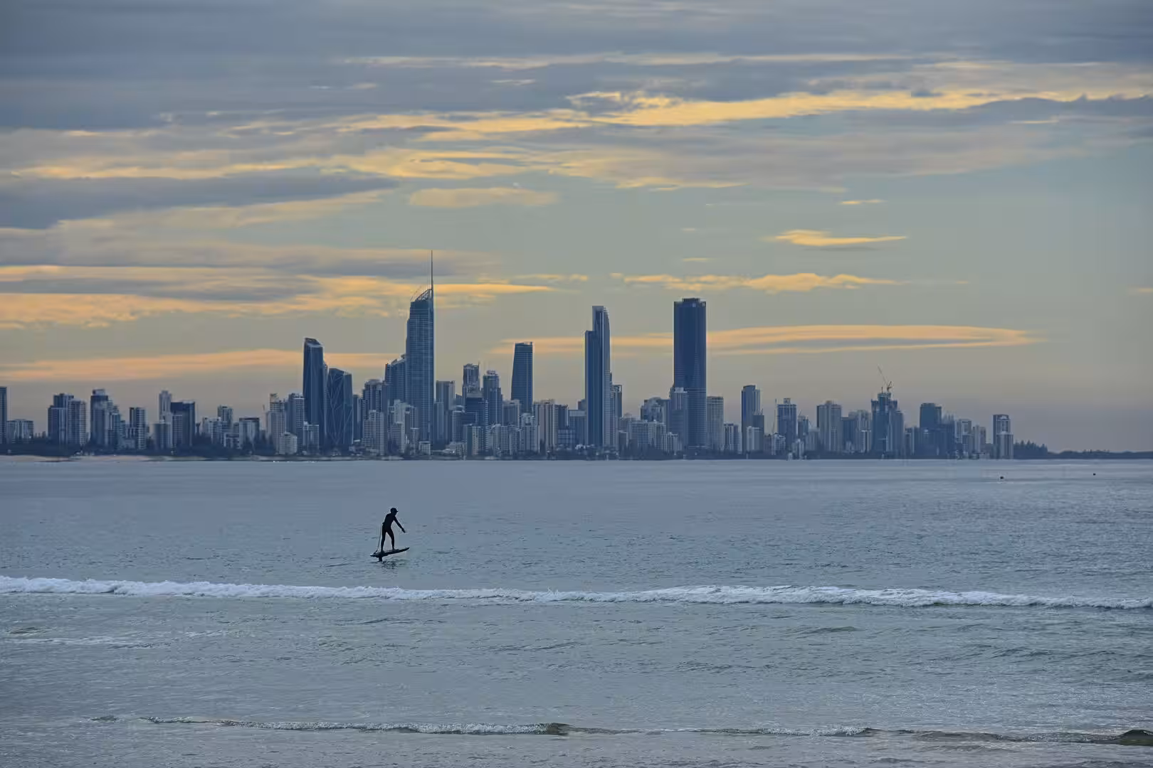 A dark paddleboarder silhouette floats on the water before a sunset-lit city skyline.