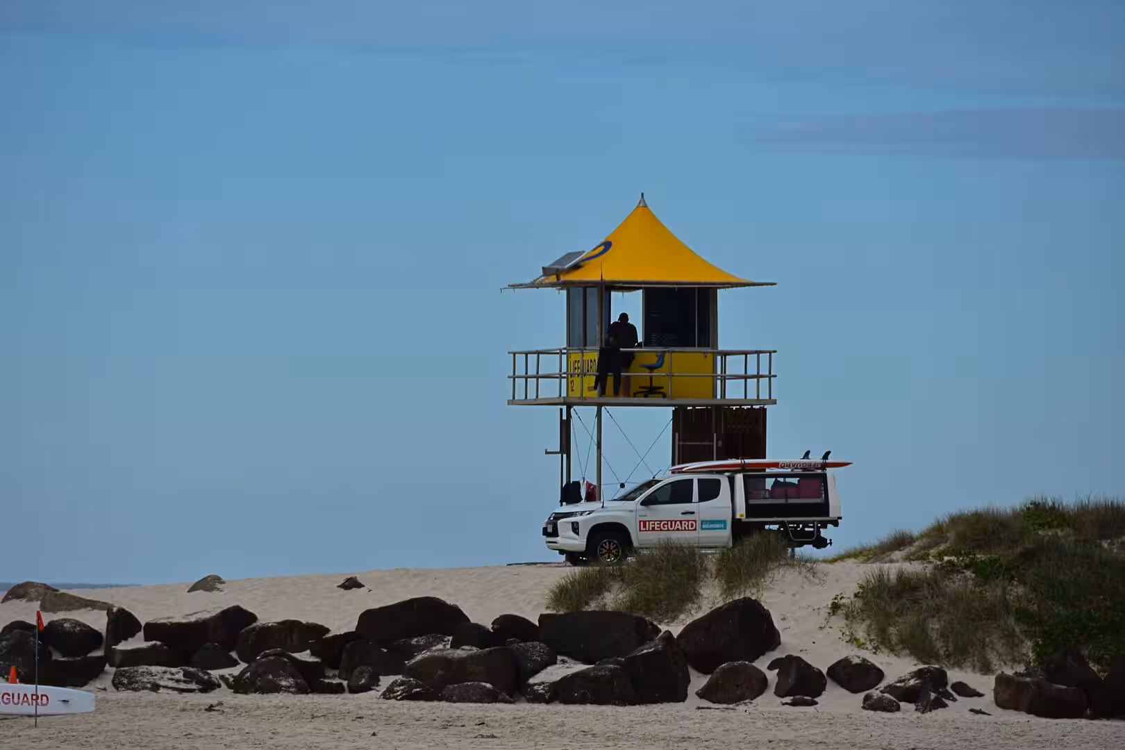 A yellow lifeguard lookout tower rises from a dune with a white patrol truck and surfboards nearby.