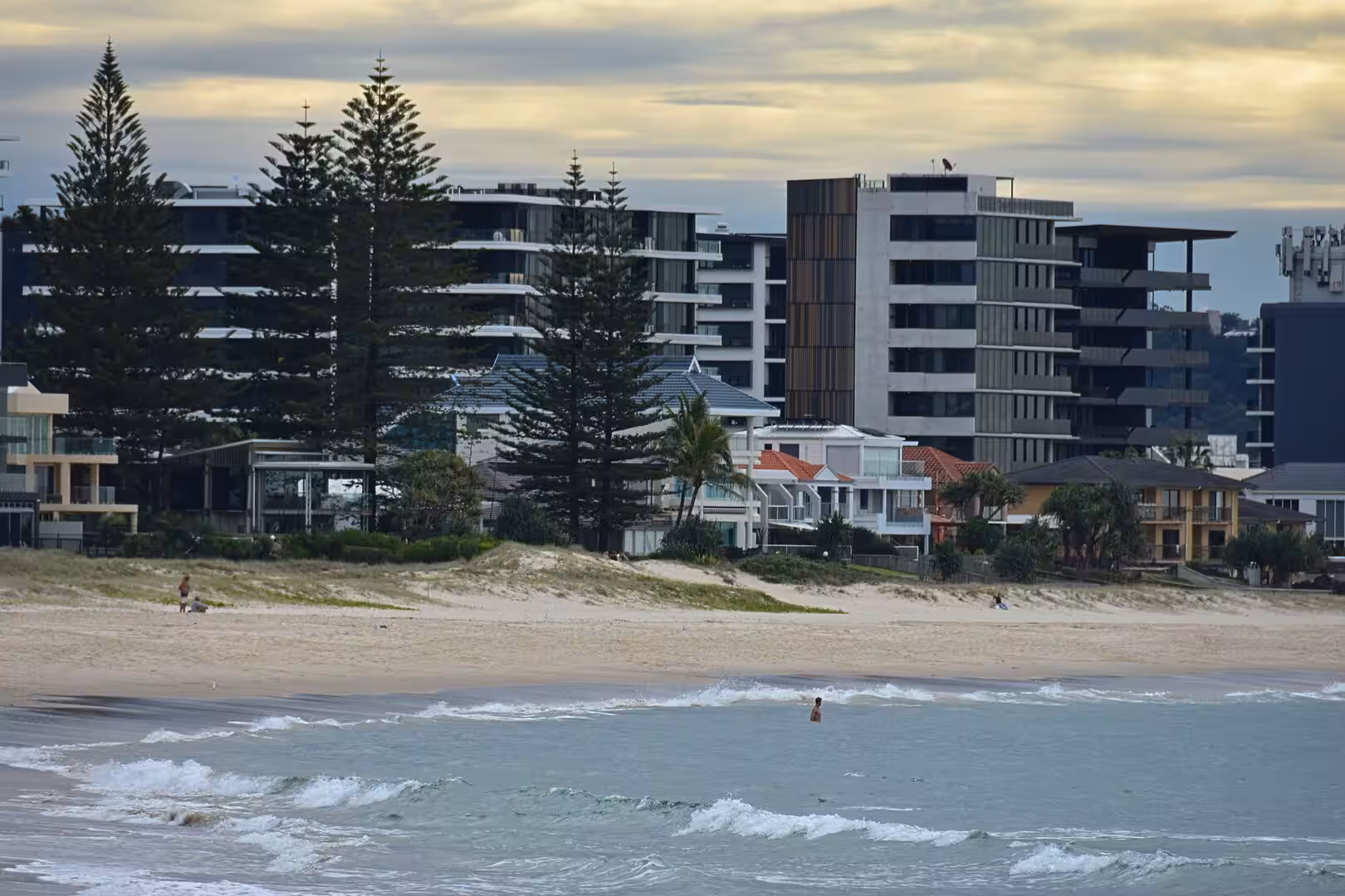 A wide sandy beach meets rolling surf, backed by tall apartment blocks and pines.