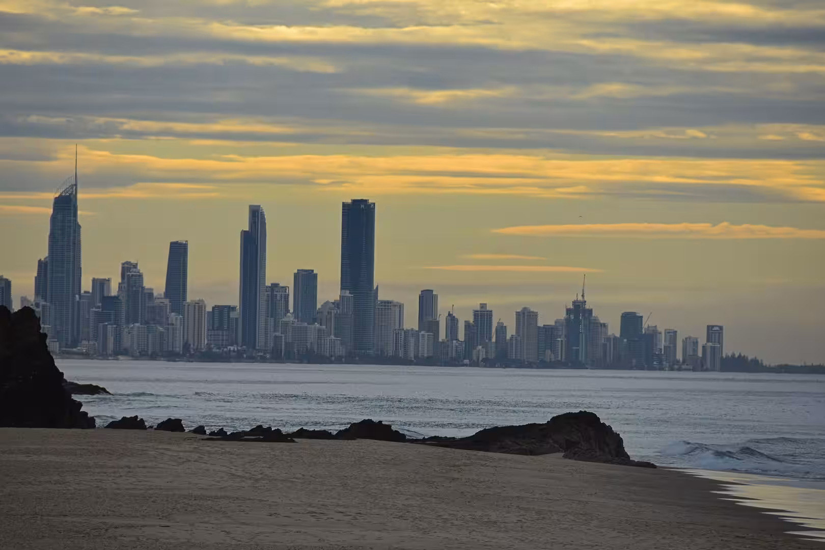 A skyline of sunlit skyscrapers rises across the water from a sandy beach.