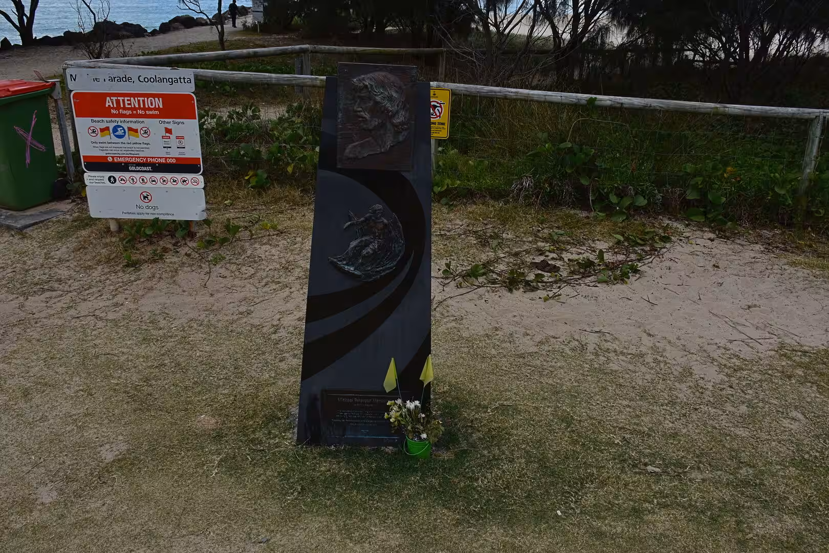 A tall dark stone memorial with flowers stands by the beach.