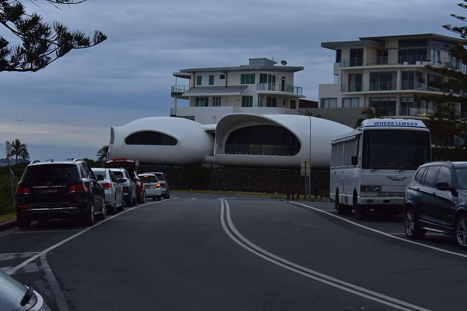 A striking white building with organic pod shapes rises above a street lined with parked cars.