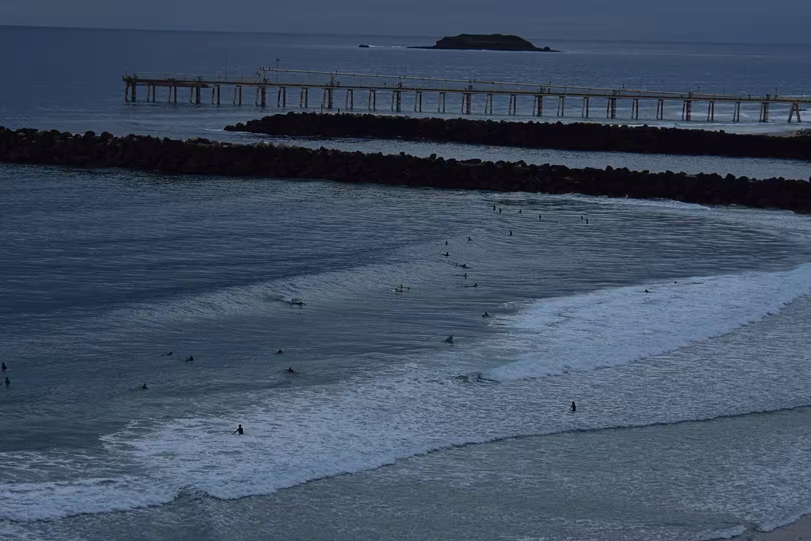 Surfers dot the rolling waves as a wooden pier and rock breakwater stretch across the dark water.